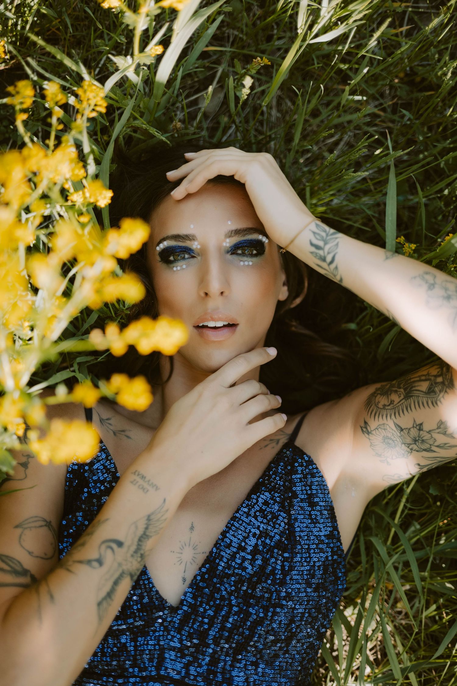 Close-up portrait with glitter makeup and tattoos among yellow flowers in garden setting.