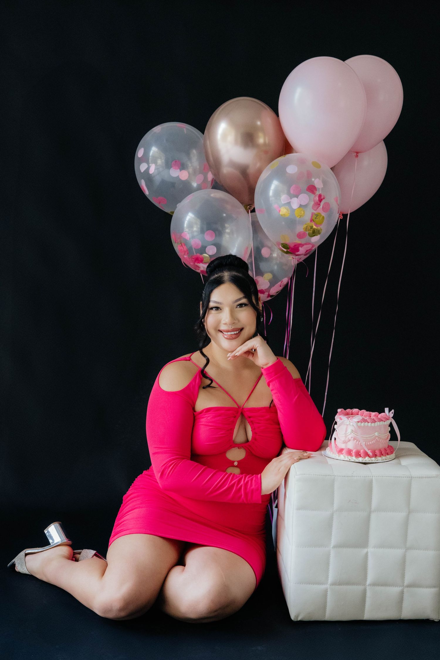 Portrait with pink and gold balloons next to white cube and small cake.