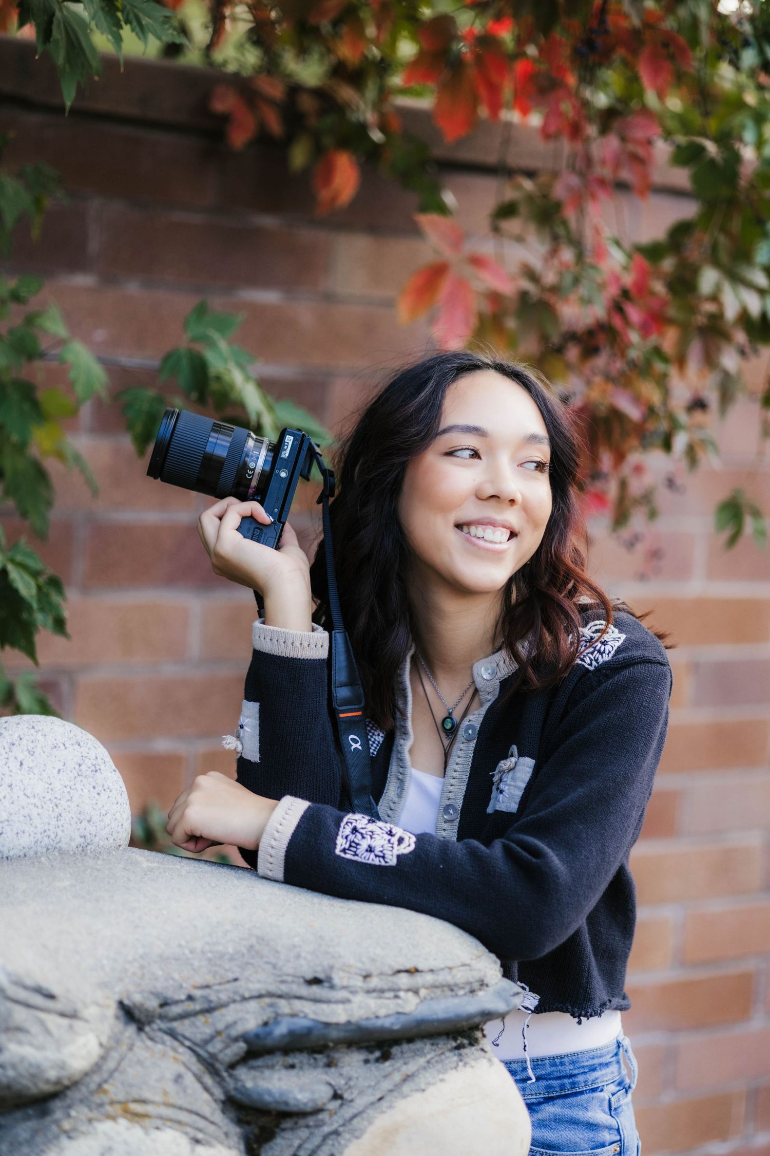 Photographer with camera leans on stone wall with autumn foliage in background.