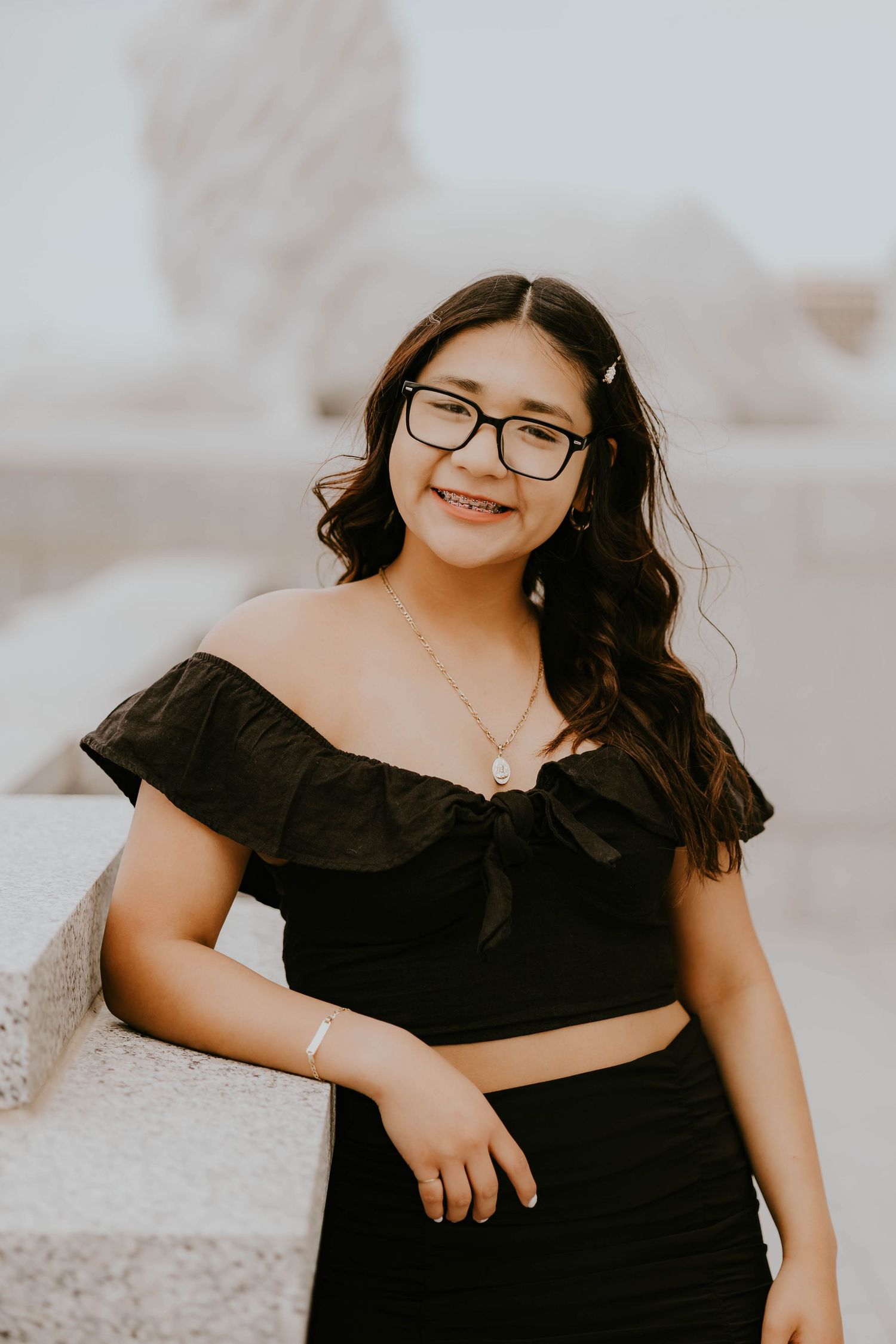 A graduate in a black off-shoulder top and glasses poses for a portrait outdoors.