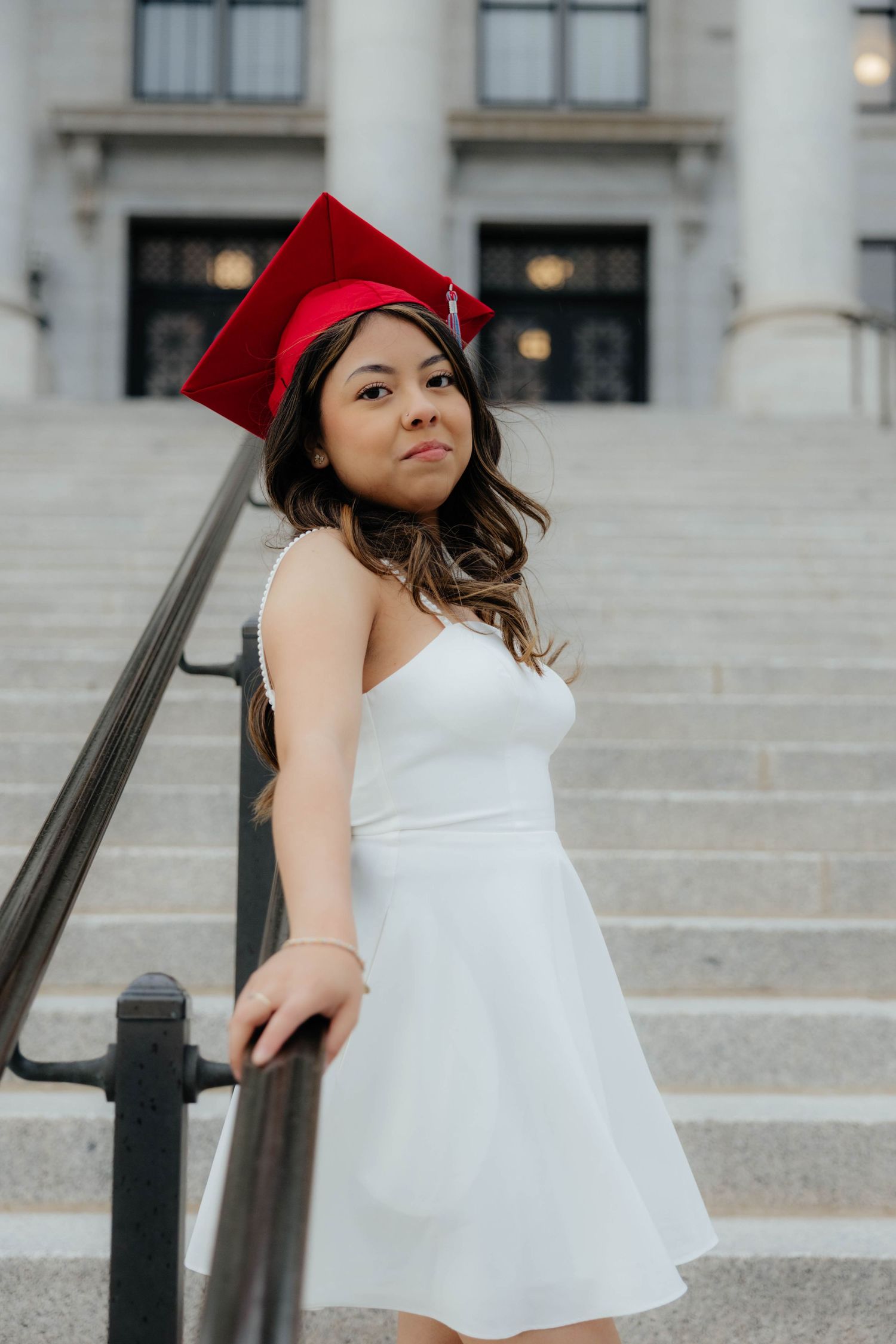 Graduate in white dress poses with red cap on stone steps of academic building.