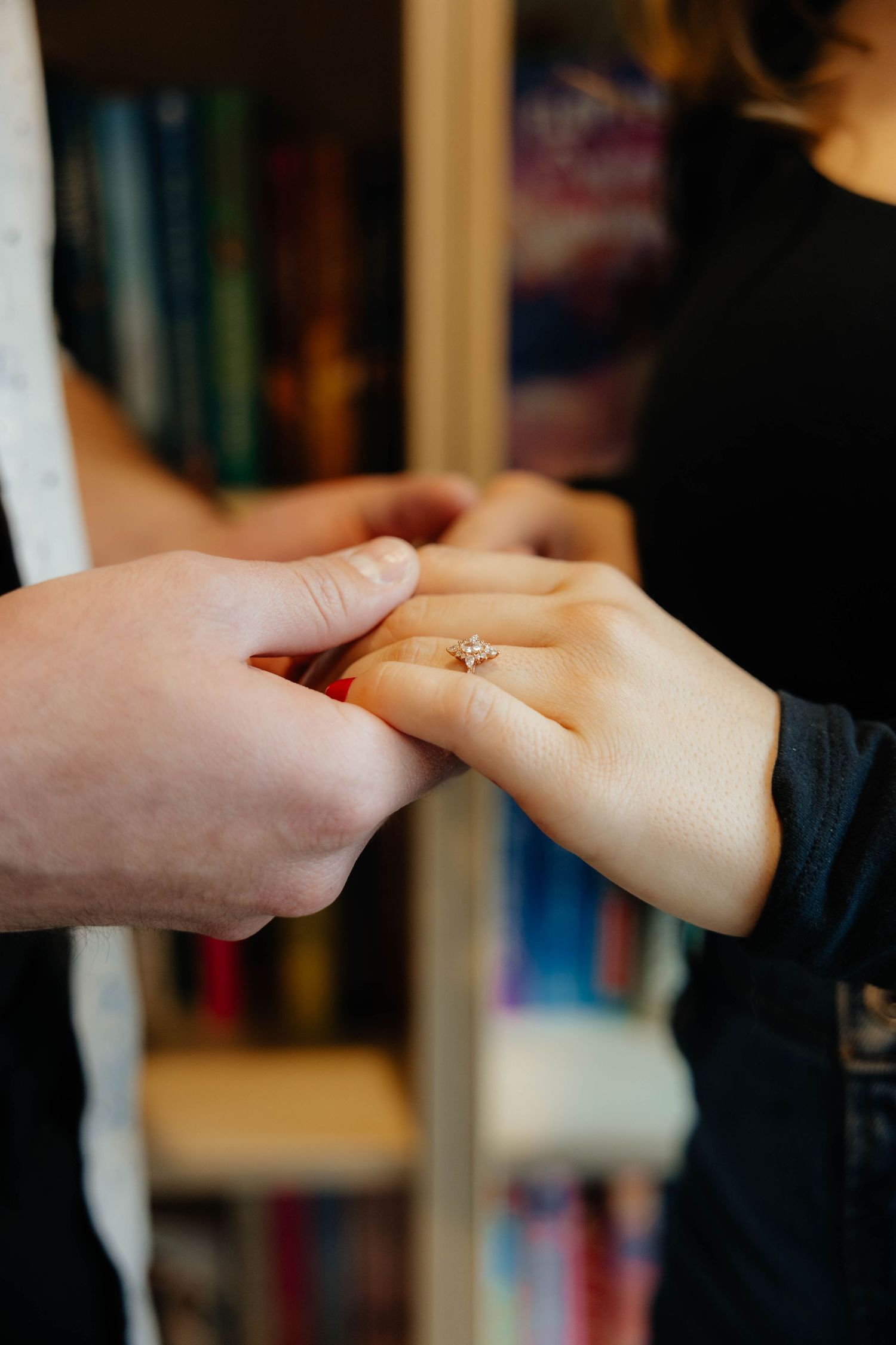 Two hands clasped together in a romantic gesture against a blurred bookshelf background.