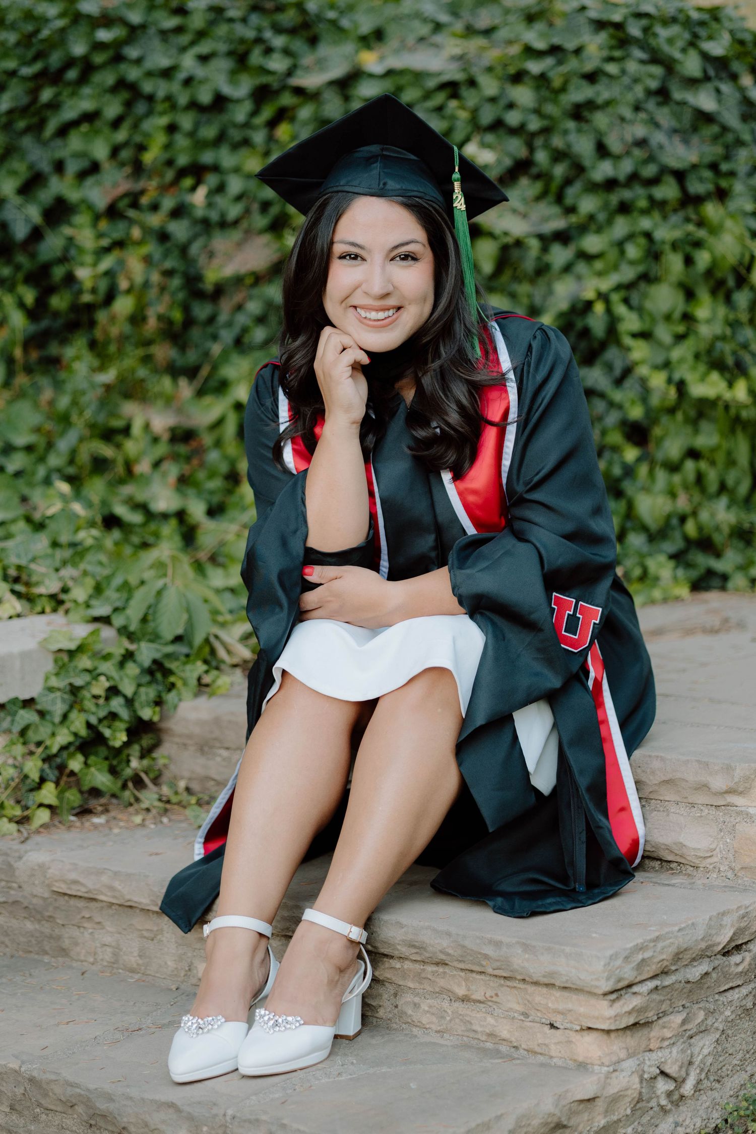 Graduate in dark cap and gown sitting on stone steps with white dress and heels.