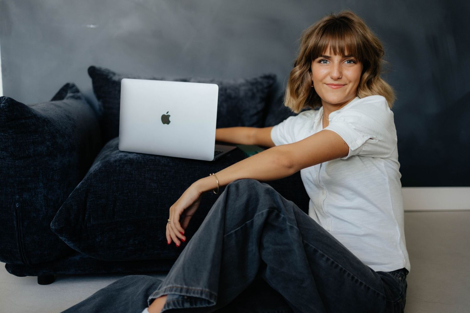 Casual portrait of woman relaxing on dark couch with MacBook laptop.
