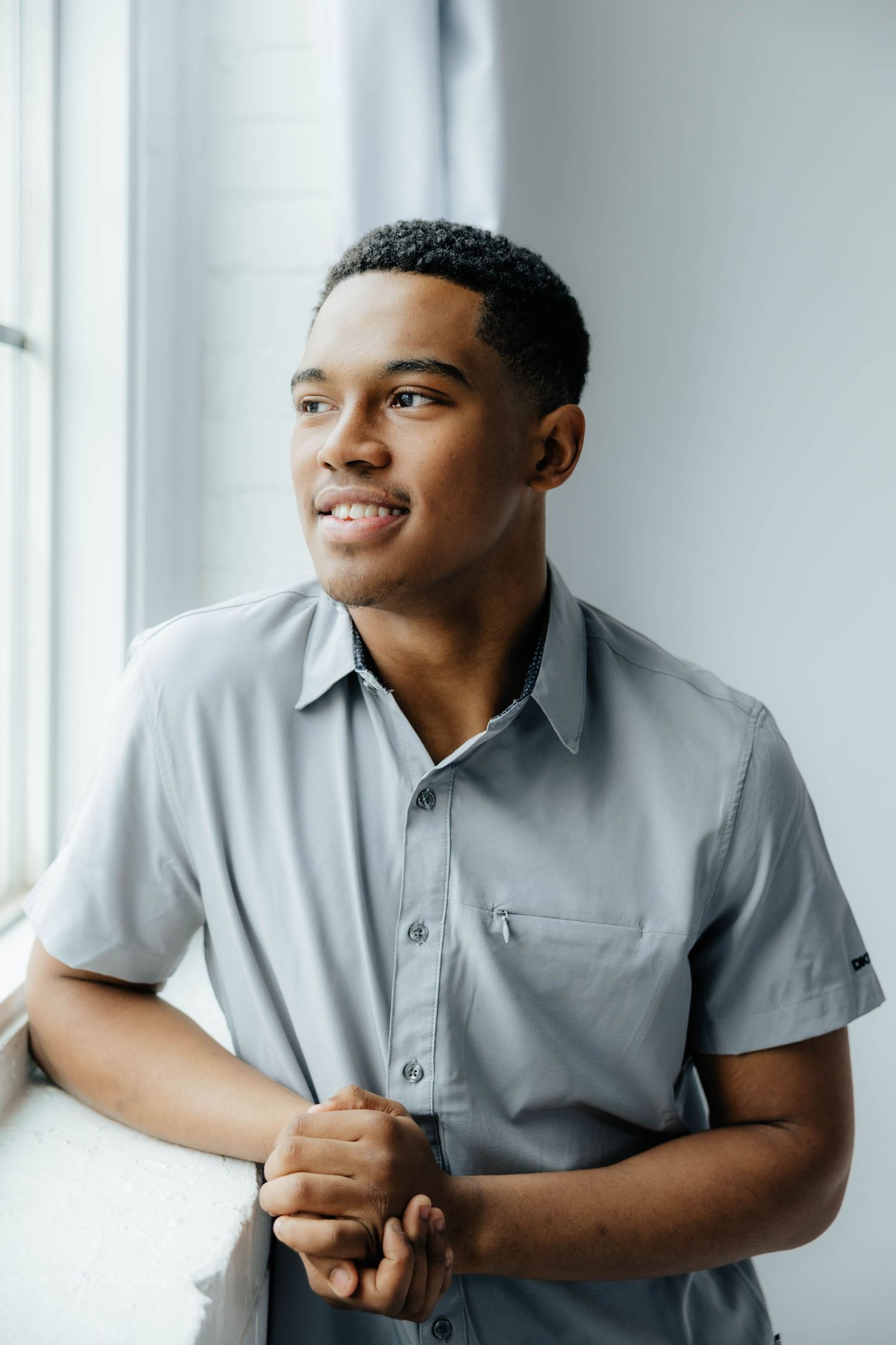 Professional in grey button-up shirt smiles while standing by window.