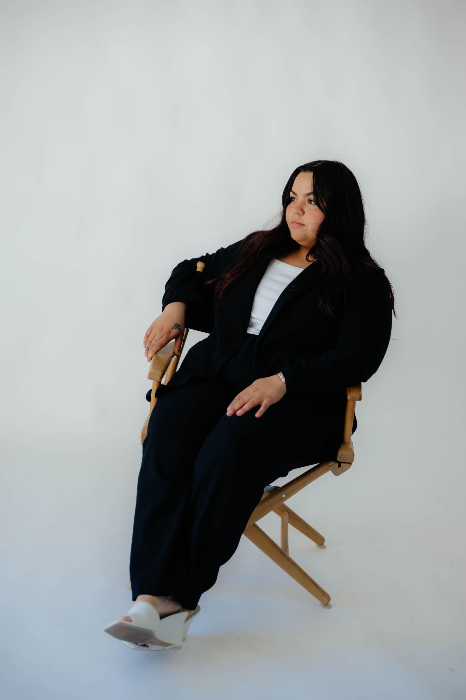 Woman in black outfit sitting on wooden chair against white studio background.
