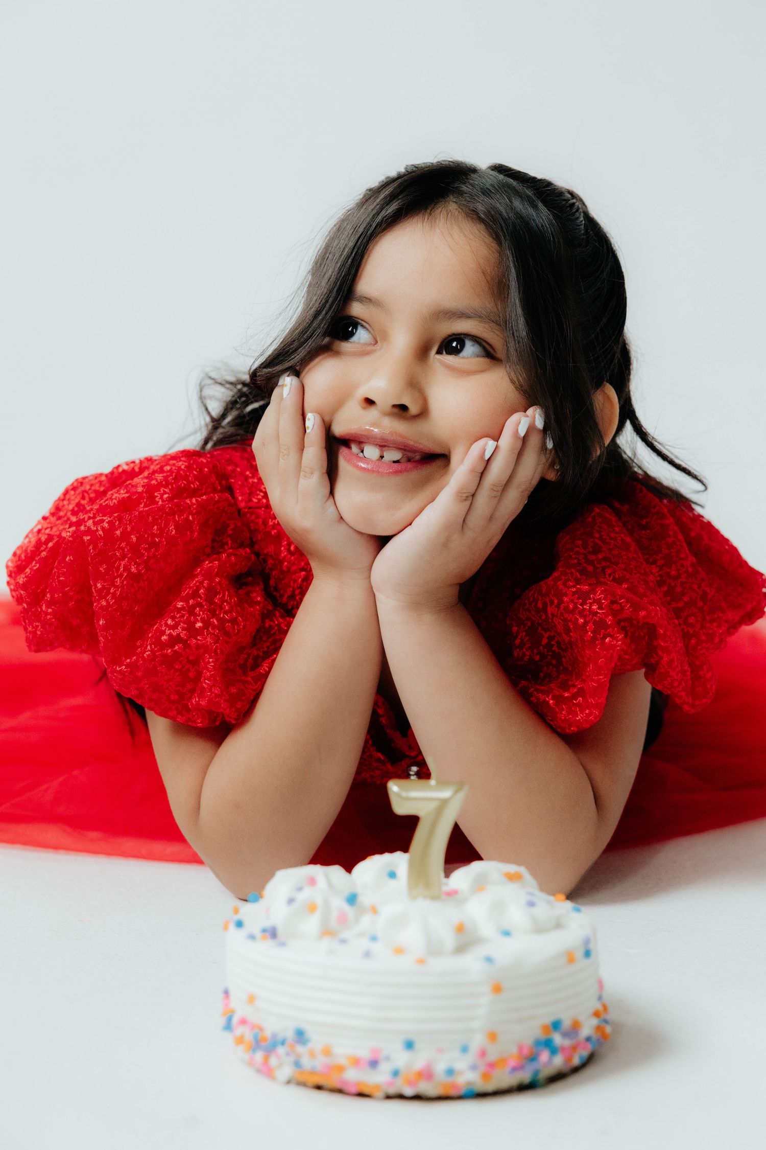 Child in red dress poses playfully with birthday cake on white background.