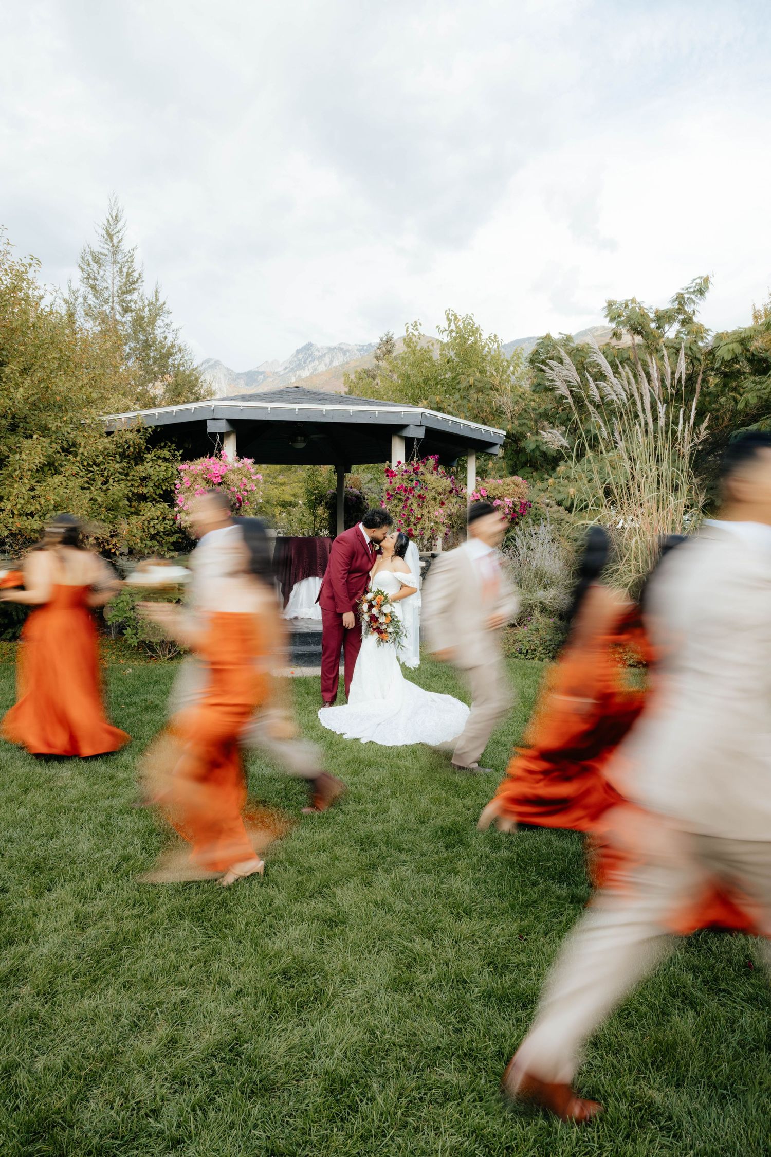 Motion-blurred wedding guests in orange and burgundy attire run across a grassy lawn during an outdoor celebration.