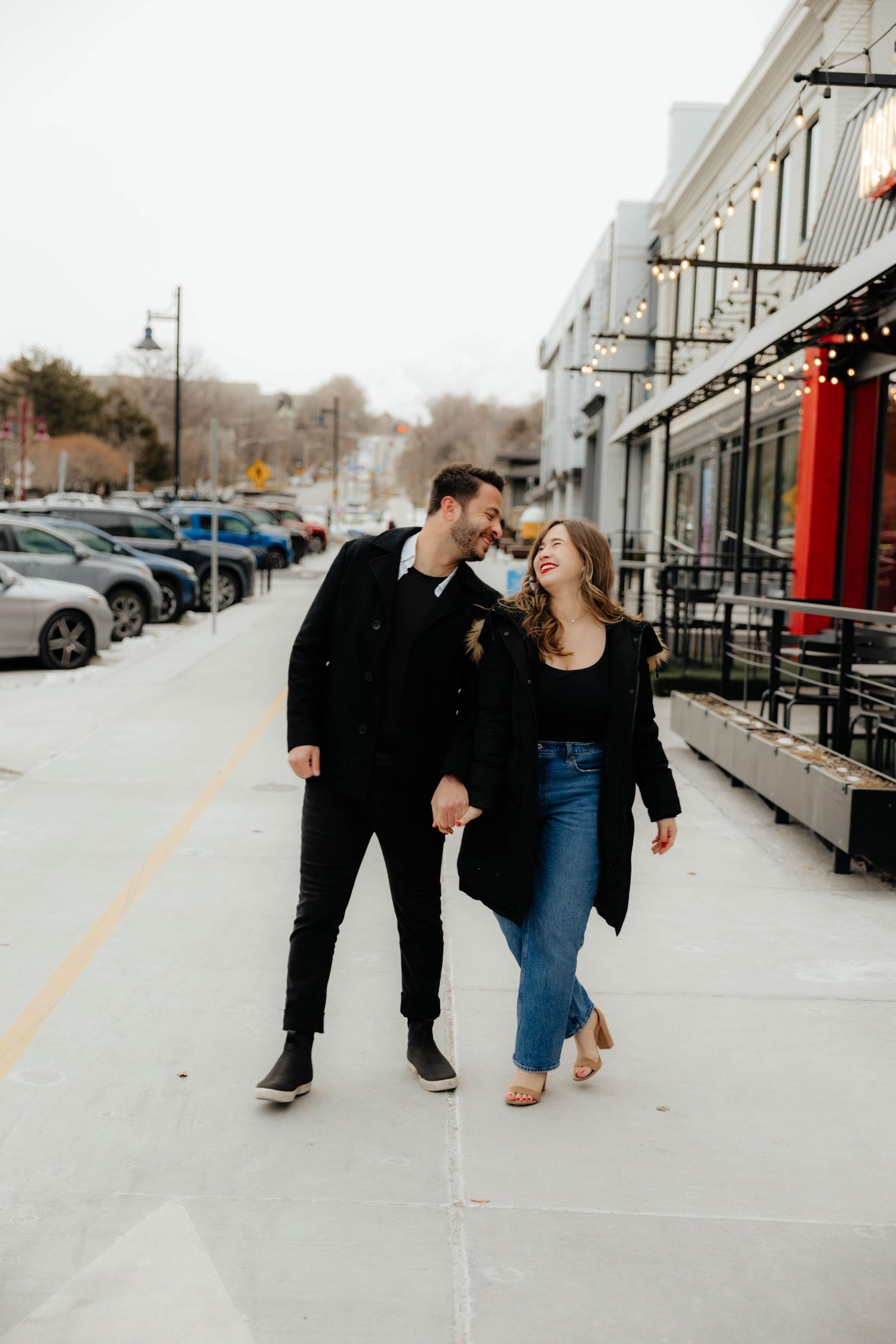 A playful couple walks and dances together in a shopping center parking lot.