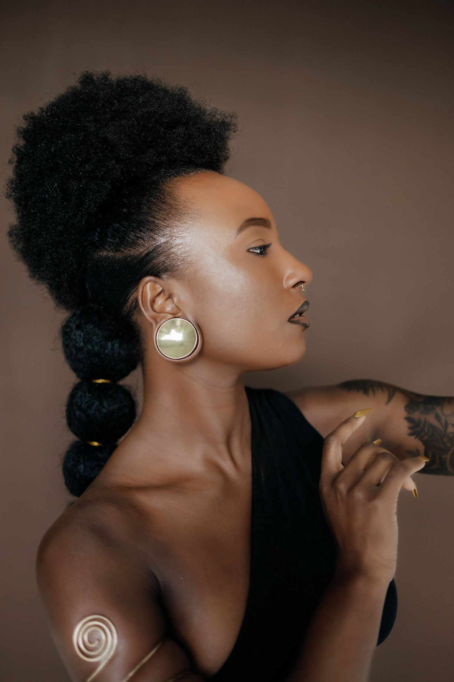 Artistic studio portrait featuring natural hair, gold earrings and elegant black attire against brown backdrop.