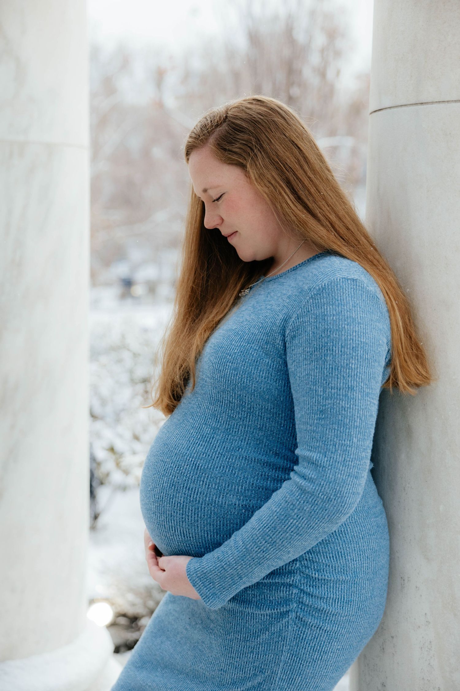 A person wearing a blue sweater poses against a white wall in a winter maternity photoshoot.