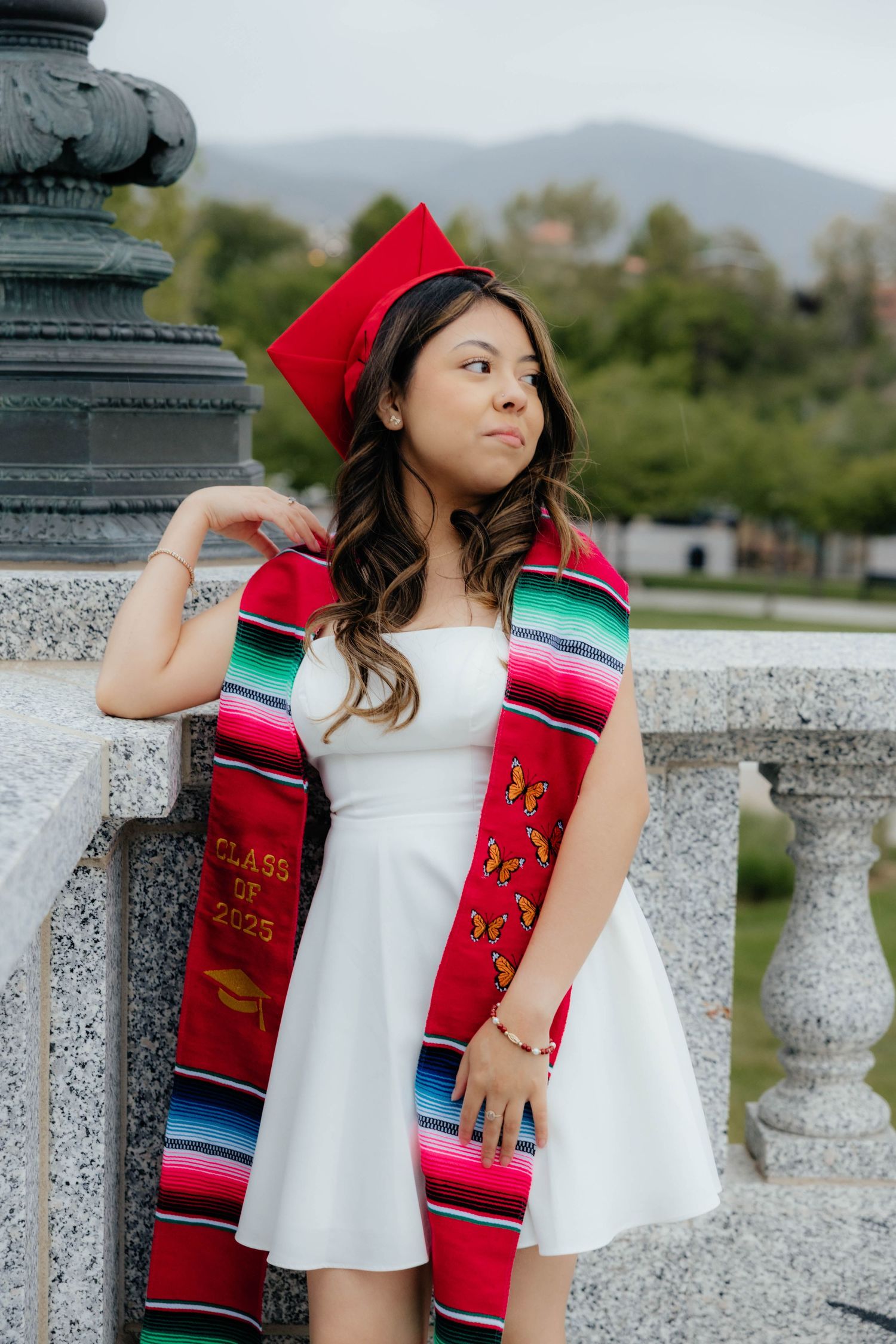 Graduate in white dress poses with colorful graduation stole against mountain backdrop.