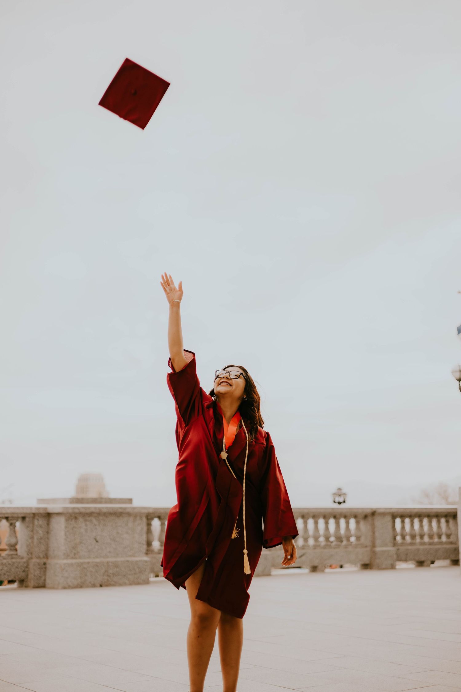 A graduate throws their red graduation cap in the air while celebrating outdoors.