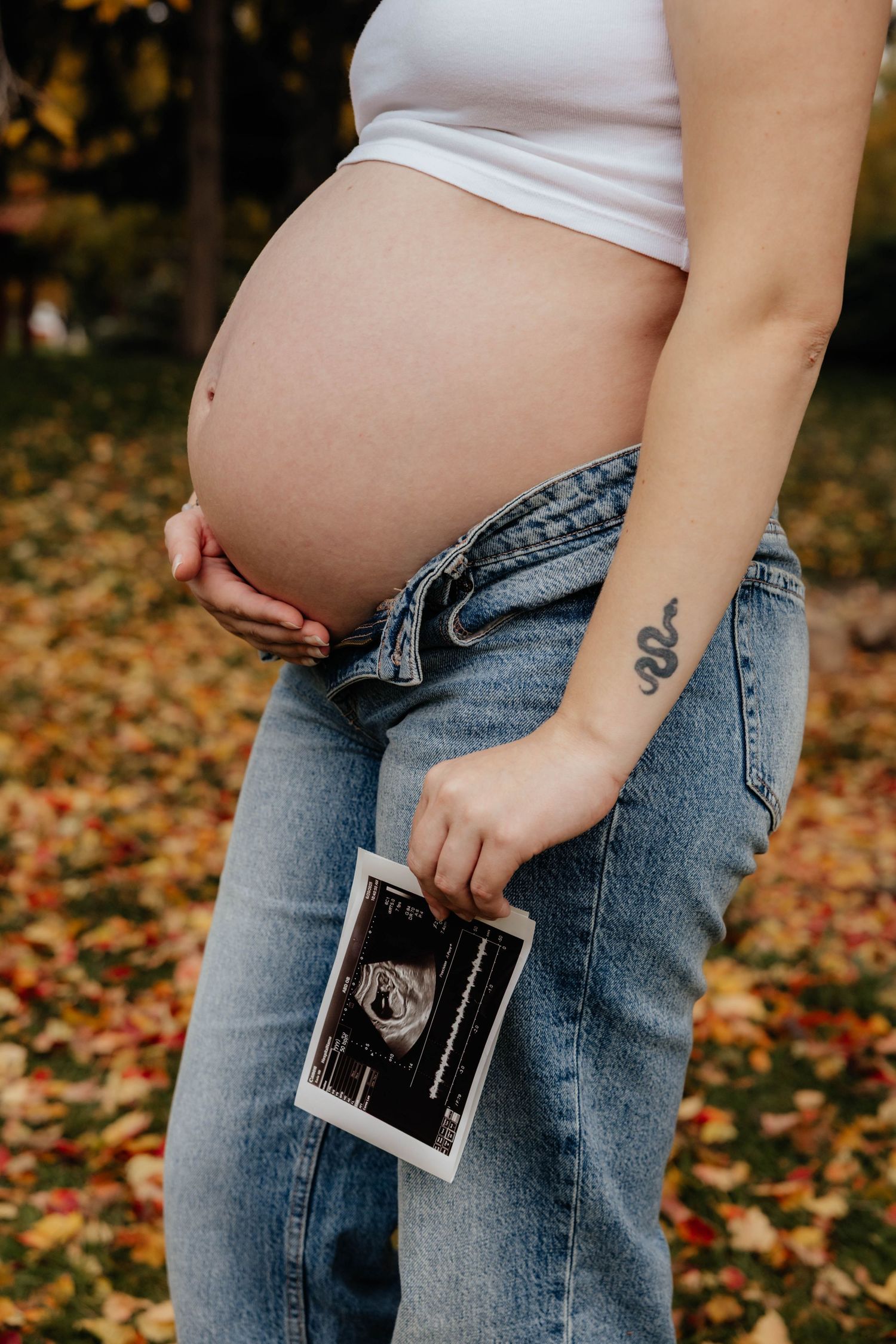 Close-up of pregnant belly with ultrasound photos against fall leaves background.