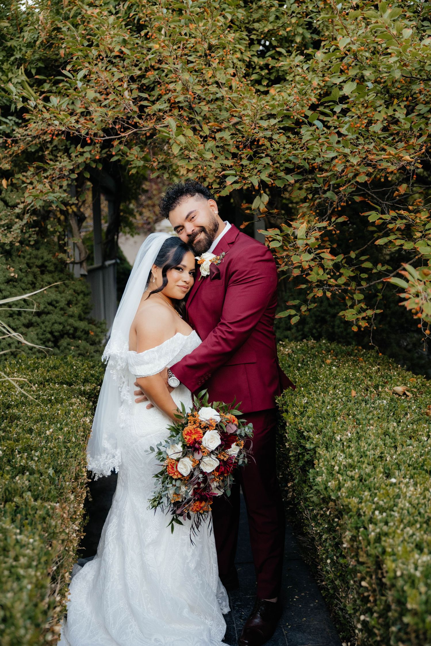 Couple embraces in garden setting with red and white floral bouquet.