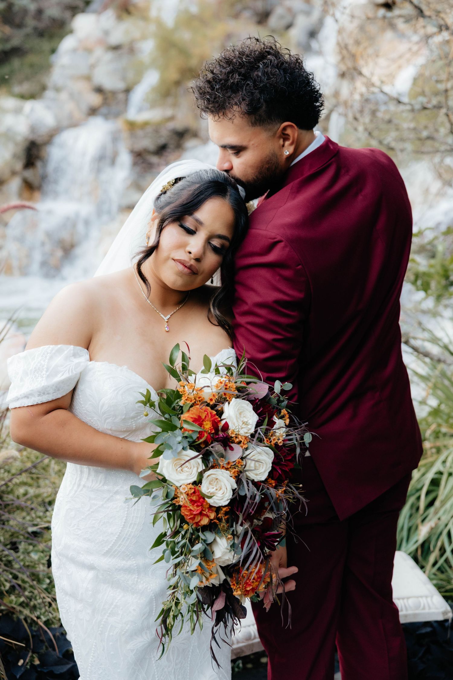 Couple embraces while holding autumn-themed wedding bouquet against rocky backdrop.
