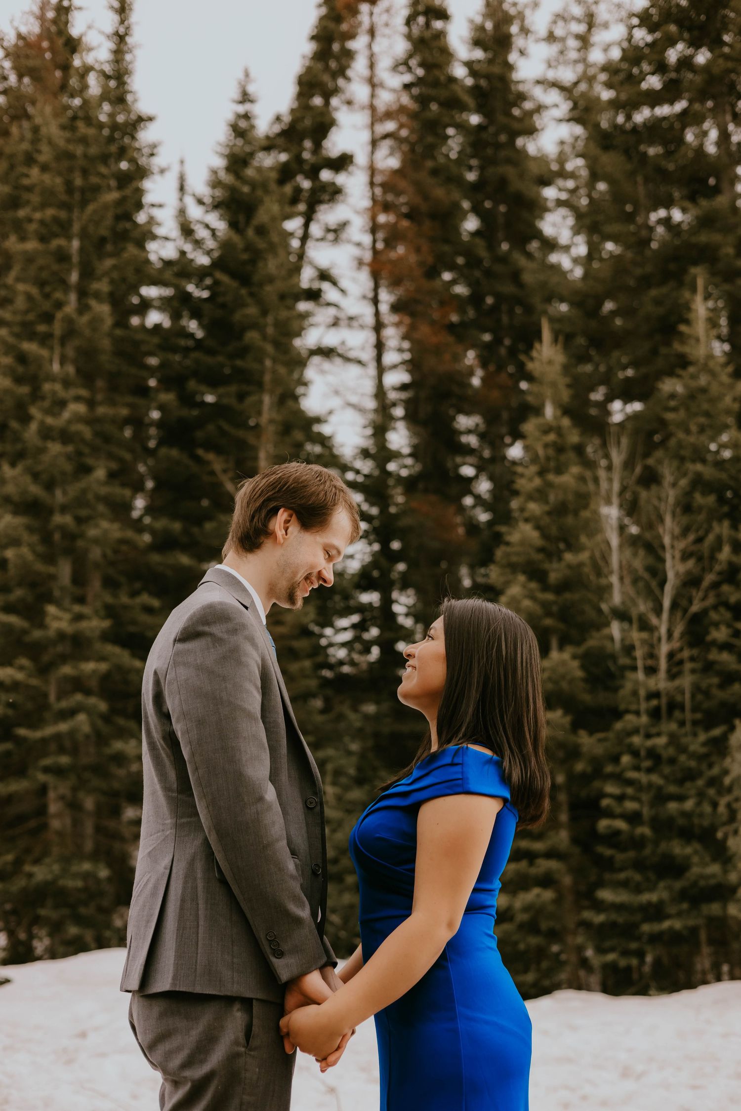 Couple in formal wear standing close together in snowy mountain setting with pine trees.