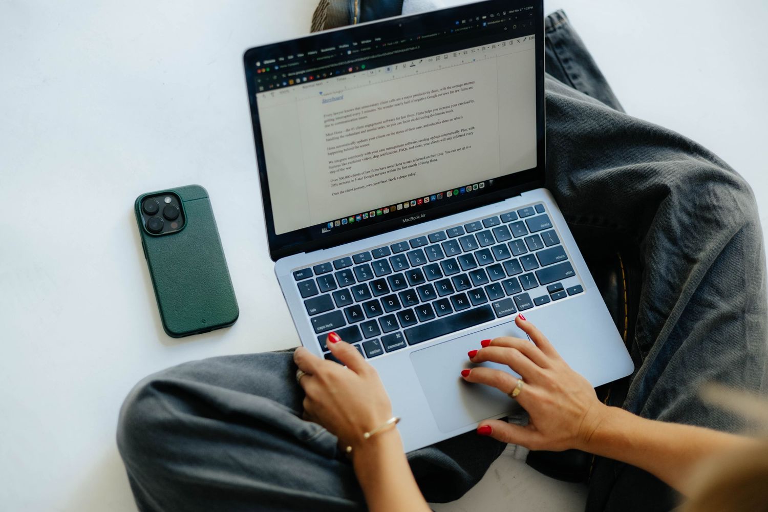 Person typing on MacBook laptop with green iPhone visible on dark surface.