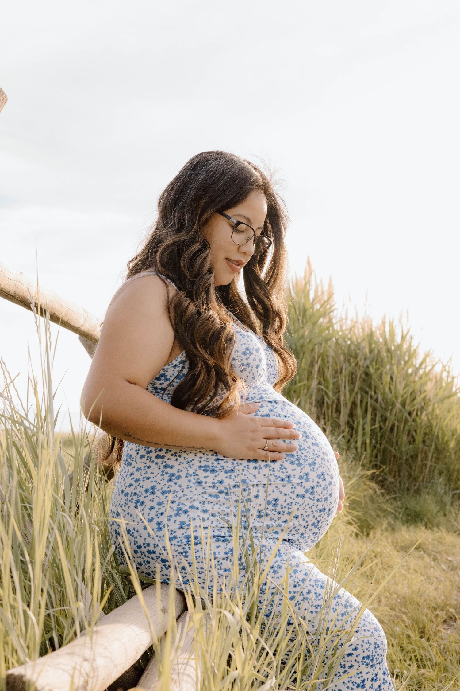 A maternity photoshoot in a field of tall grass at sunset featuring a floral blue and white dress.