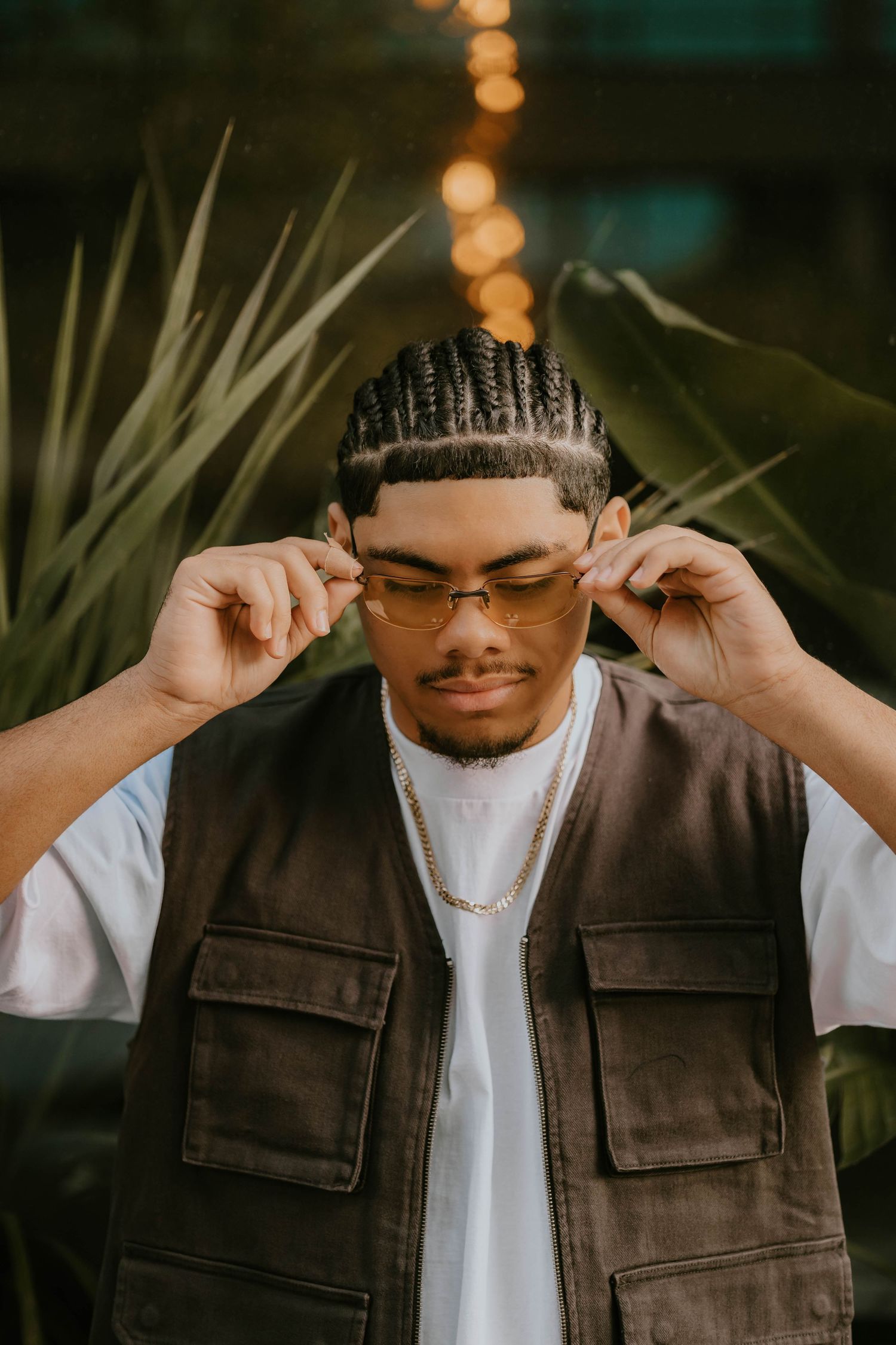 Fashion photoshoot featuring person in dark utility vest and white t-shirt adjusting sunglasses.