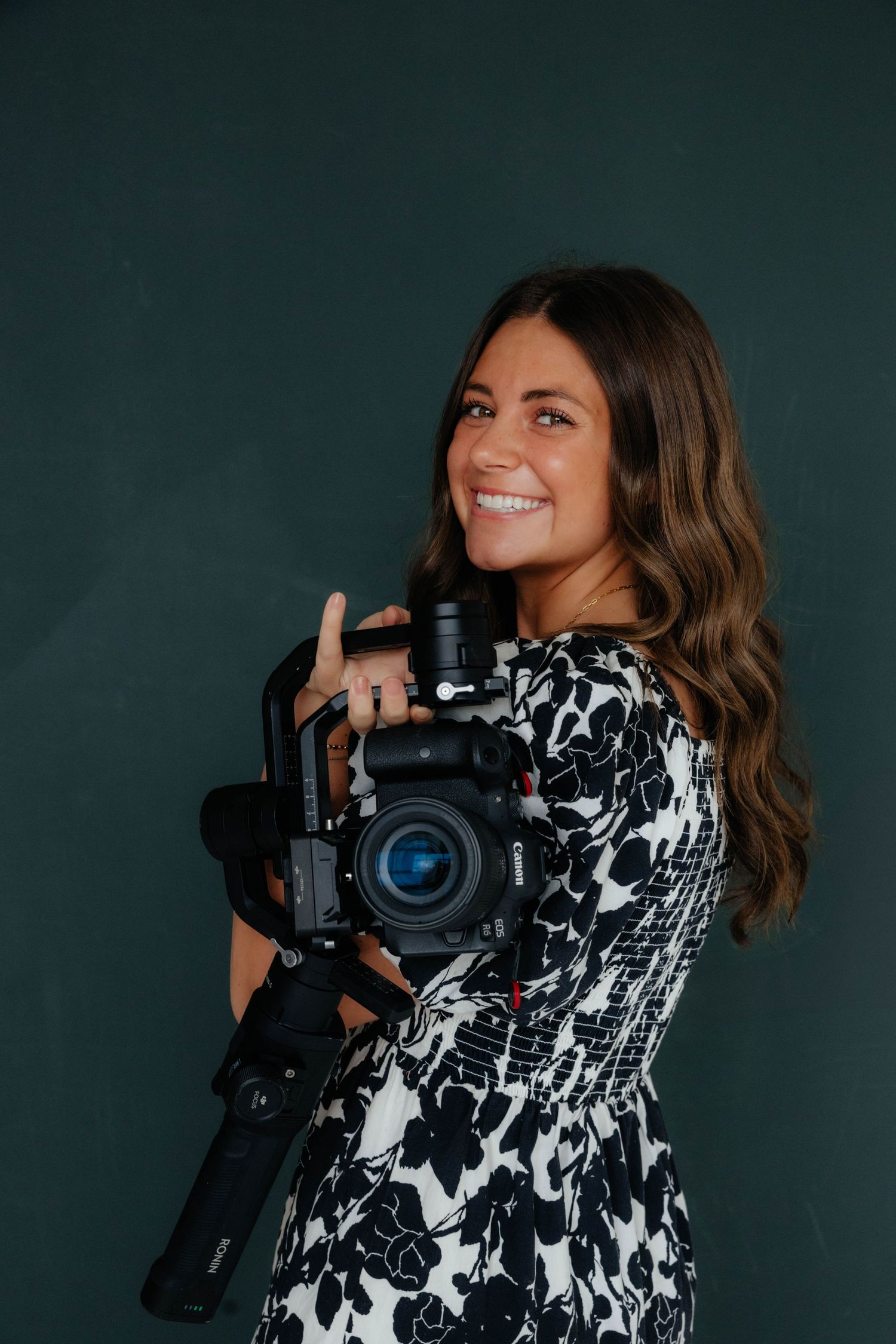 Professional photographer in black and white floral dress holding professional camera against dark green background.