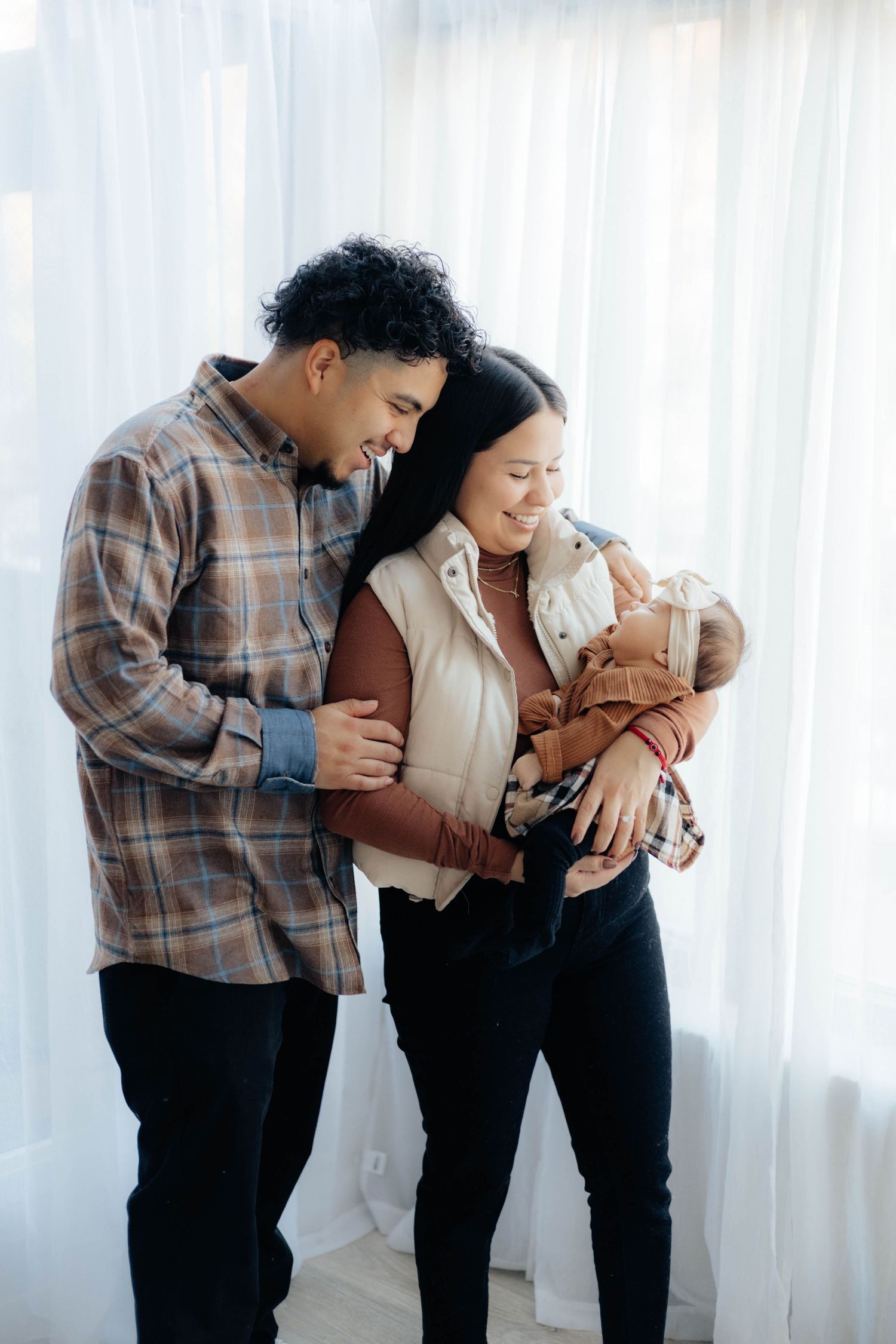 Parents share an intimate moment with their newborn in a bright, window-lit setting.