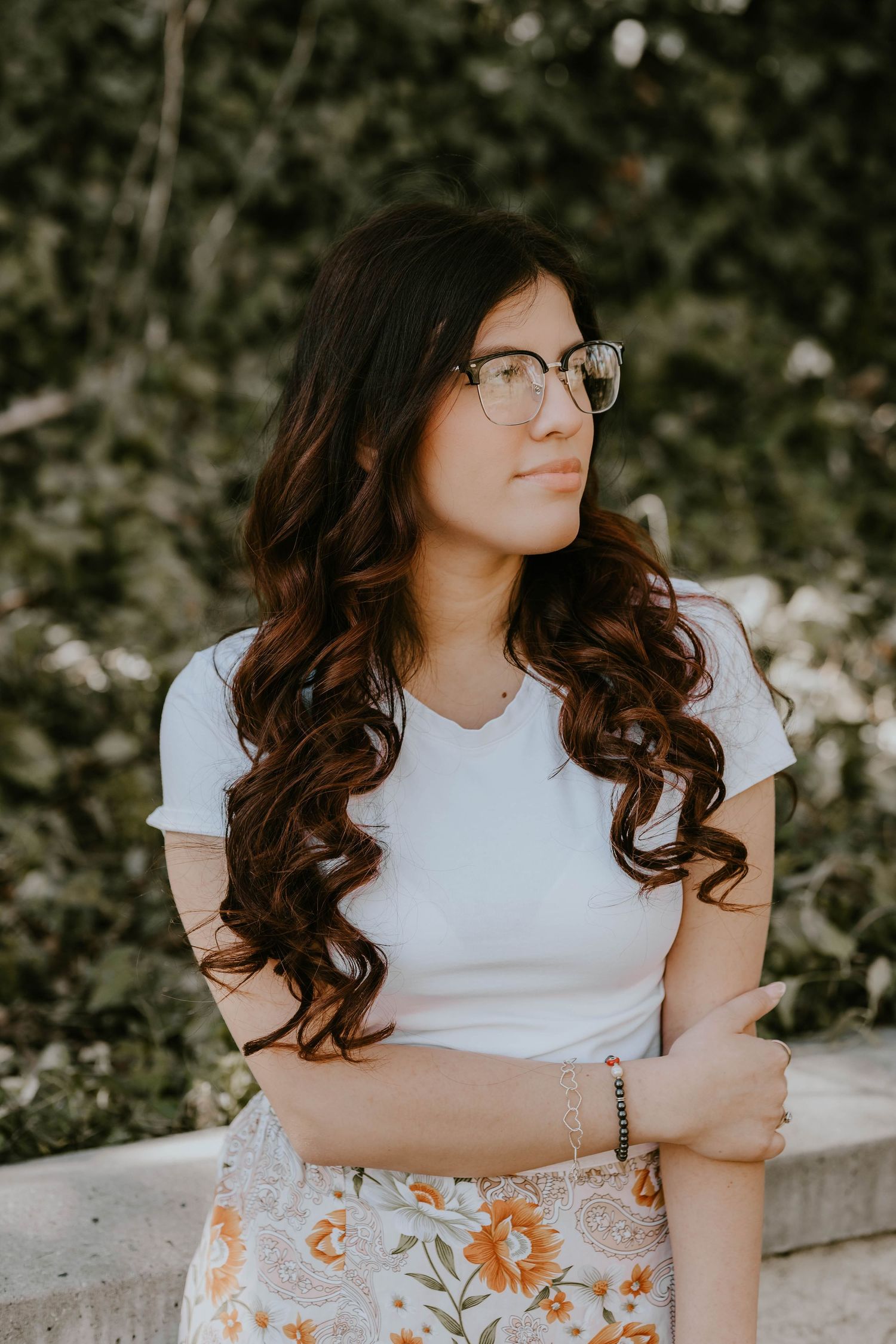 Person in white t-shirt sitting outdoors with wavy hair and glasses.