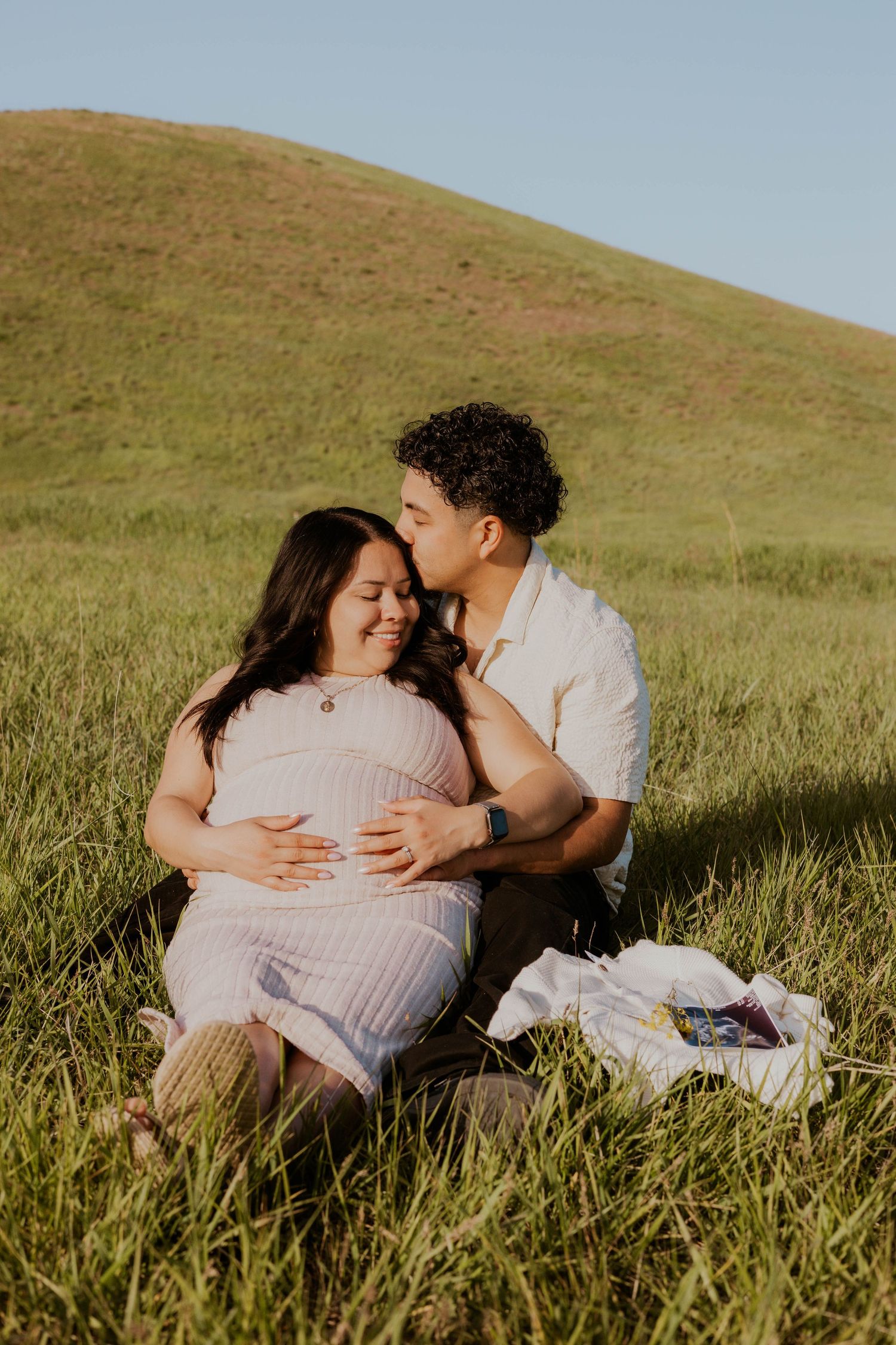 Couple sharing intimate moment while lying in tall grass during sunset maternity shoot.