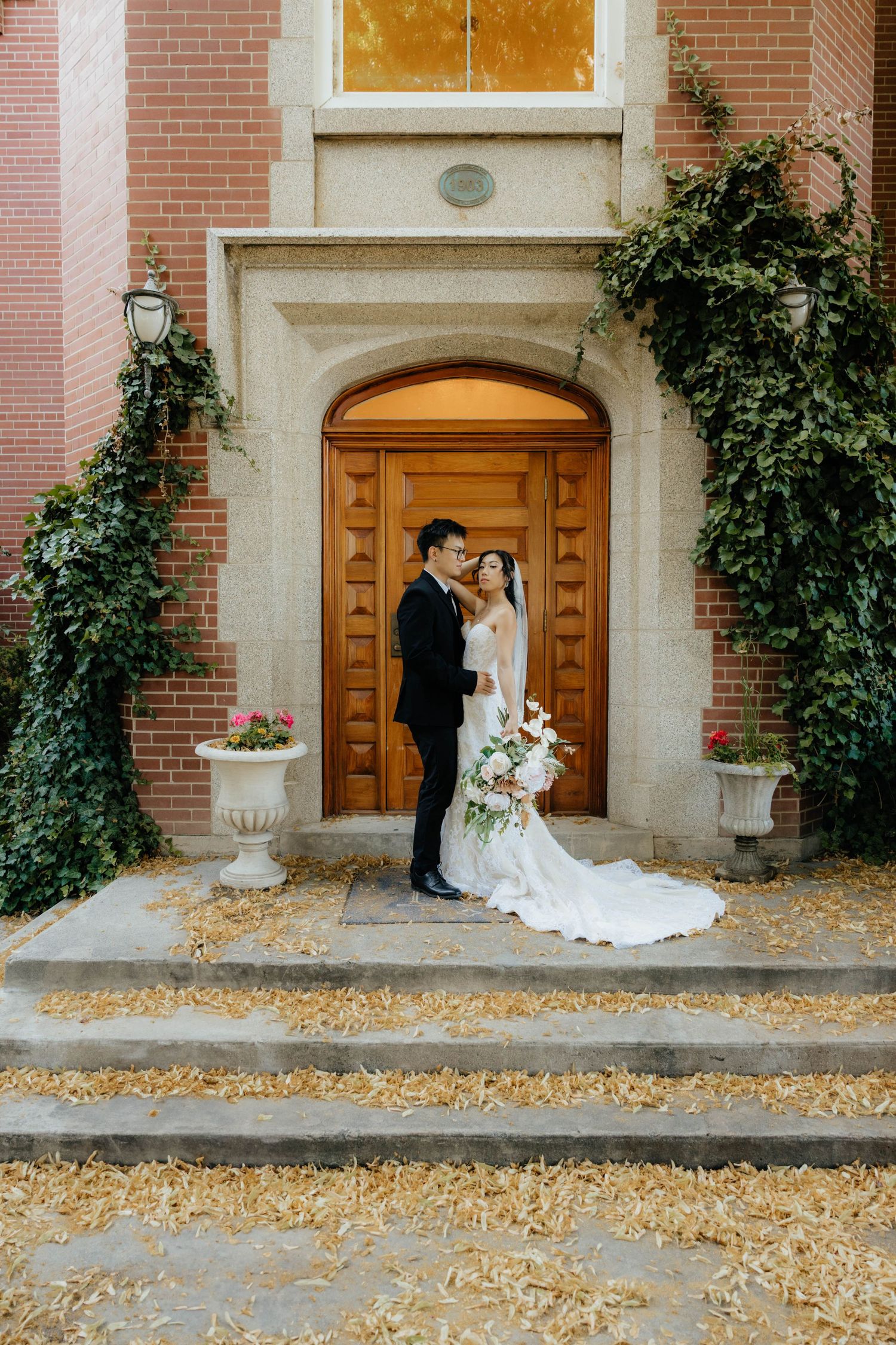 Newlyweds share intimate moment outside historic brick building with autumn leaves.