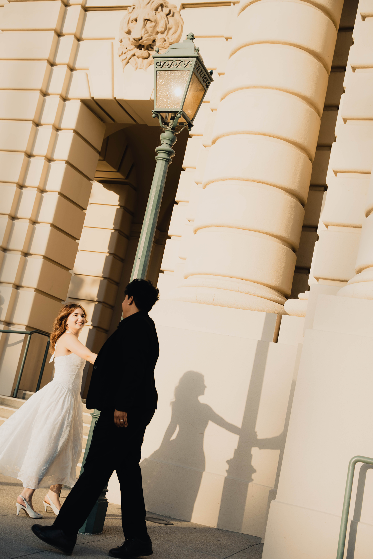 Bride and groom walk hand-in-hand past ornate building columns, their shadows cast romantically on the warm sunlit wall.