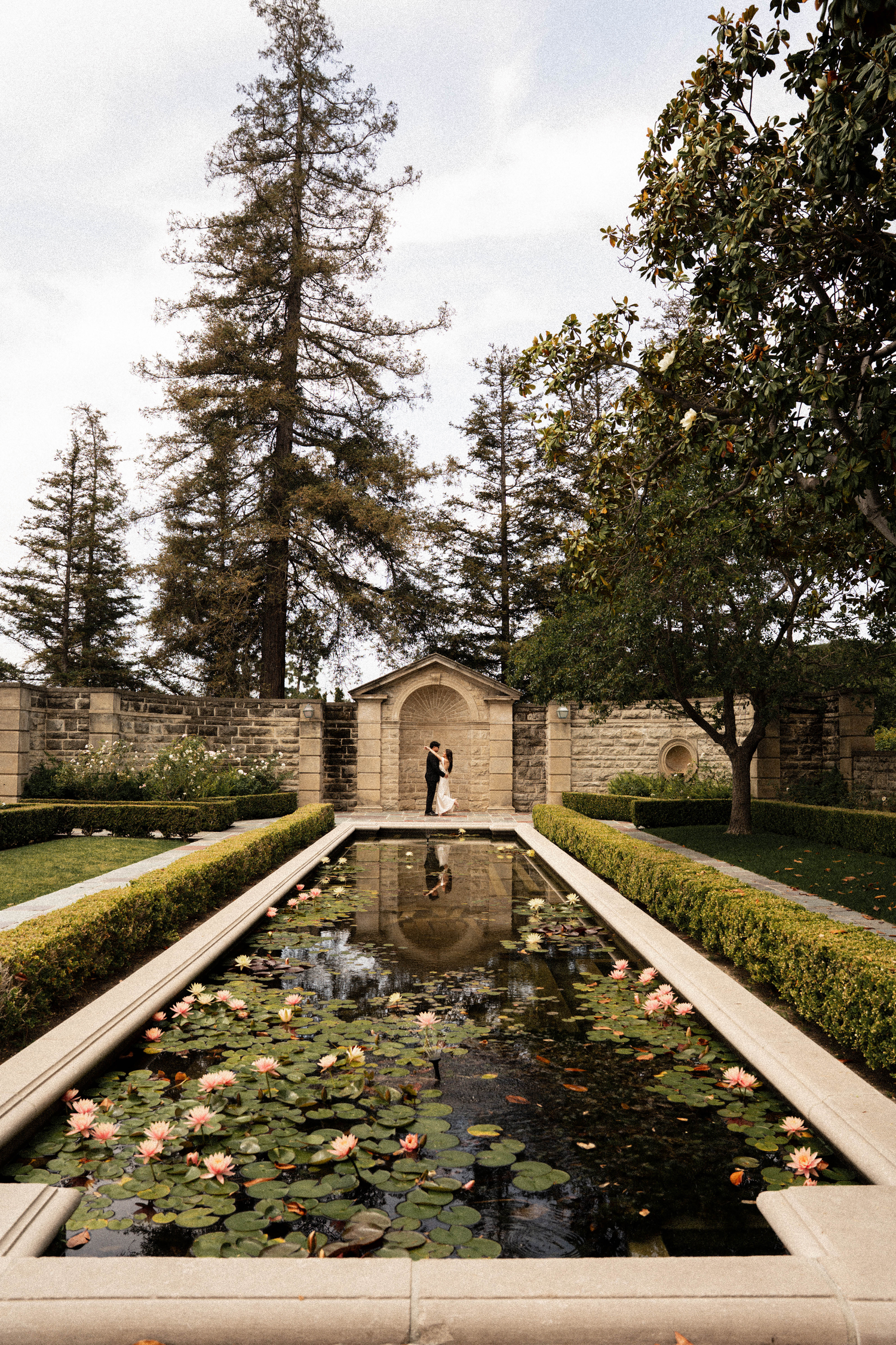 Couple embraces by a serene lily pond in a formal garden with stone archway and tall trees.