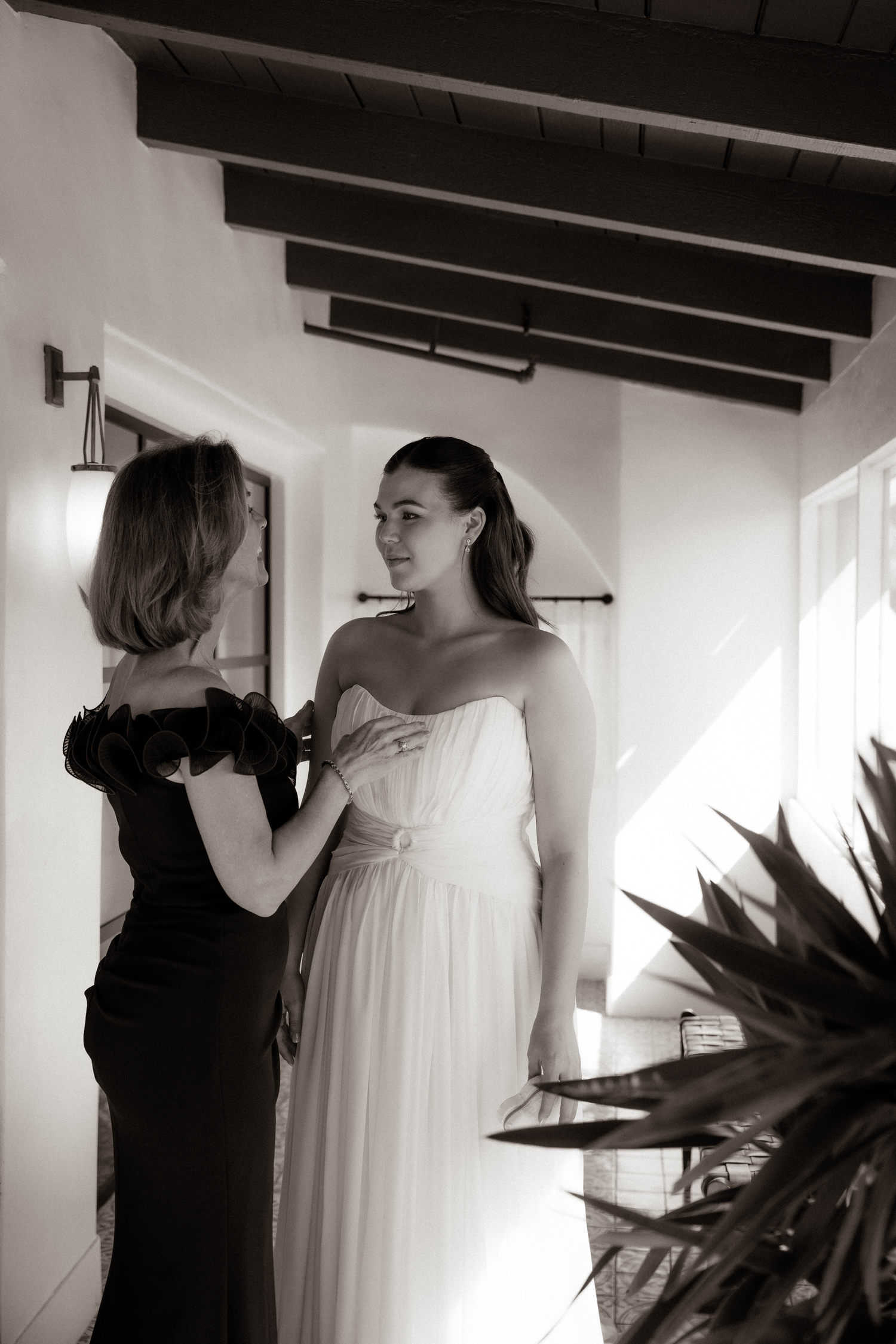 Black and white photo of a bride in a strapless gown being helped by a woman in a Spanish-style venue.