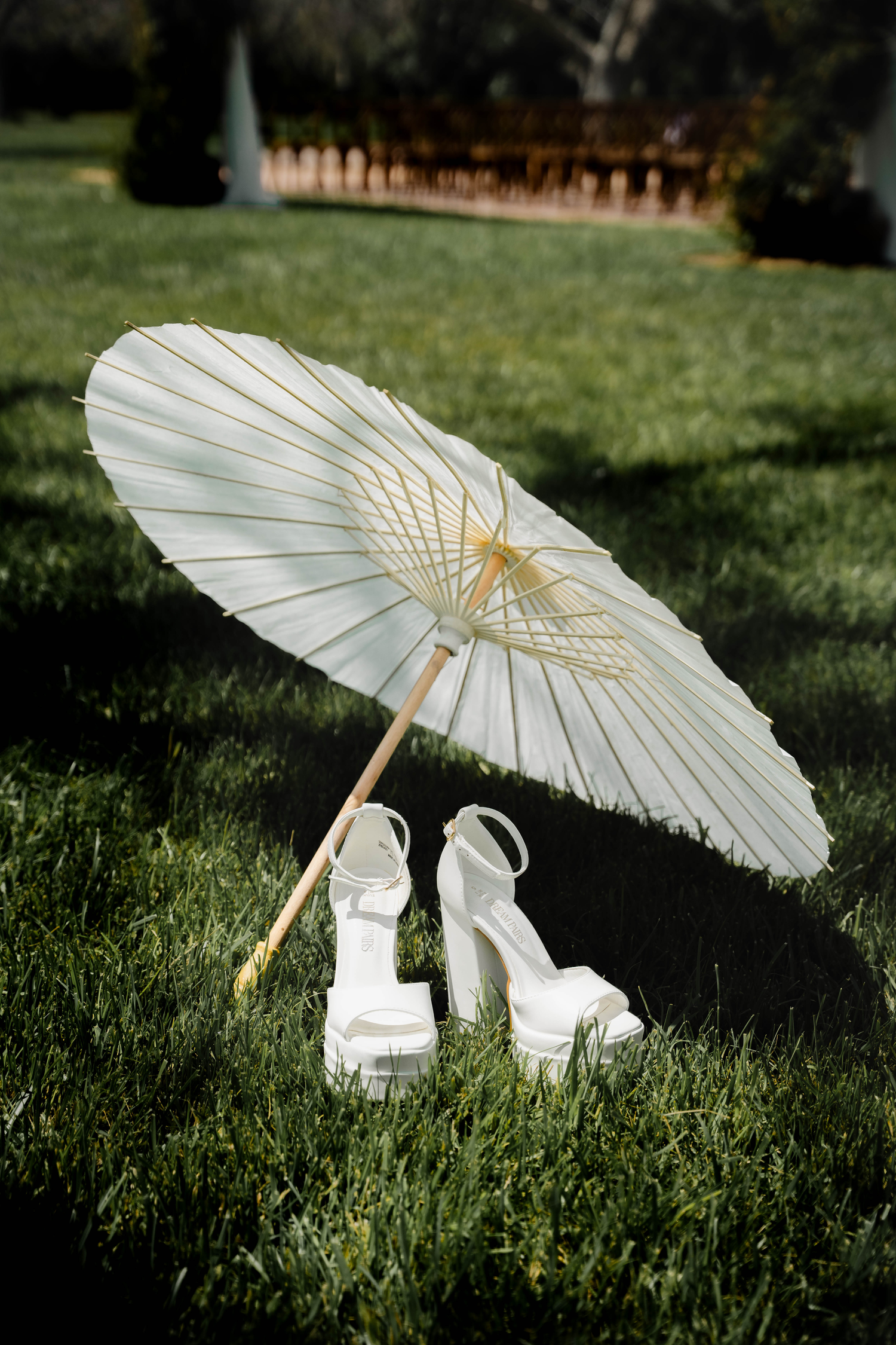 White paper parasol umbrella leaning on grass beside elegant white platform heeled bridal shoes at an outdoor wedding.