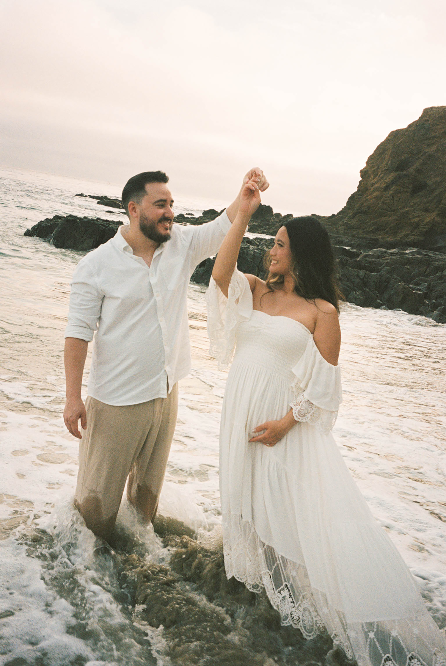 Expecting couple dancing in ocean waves at sunset, maternity beach photography session.