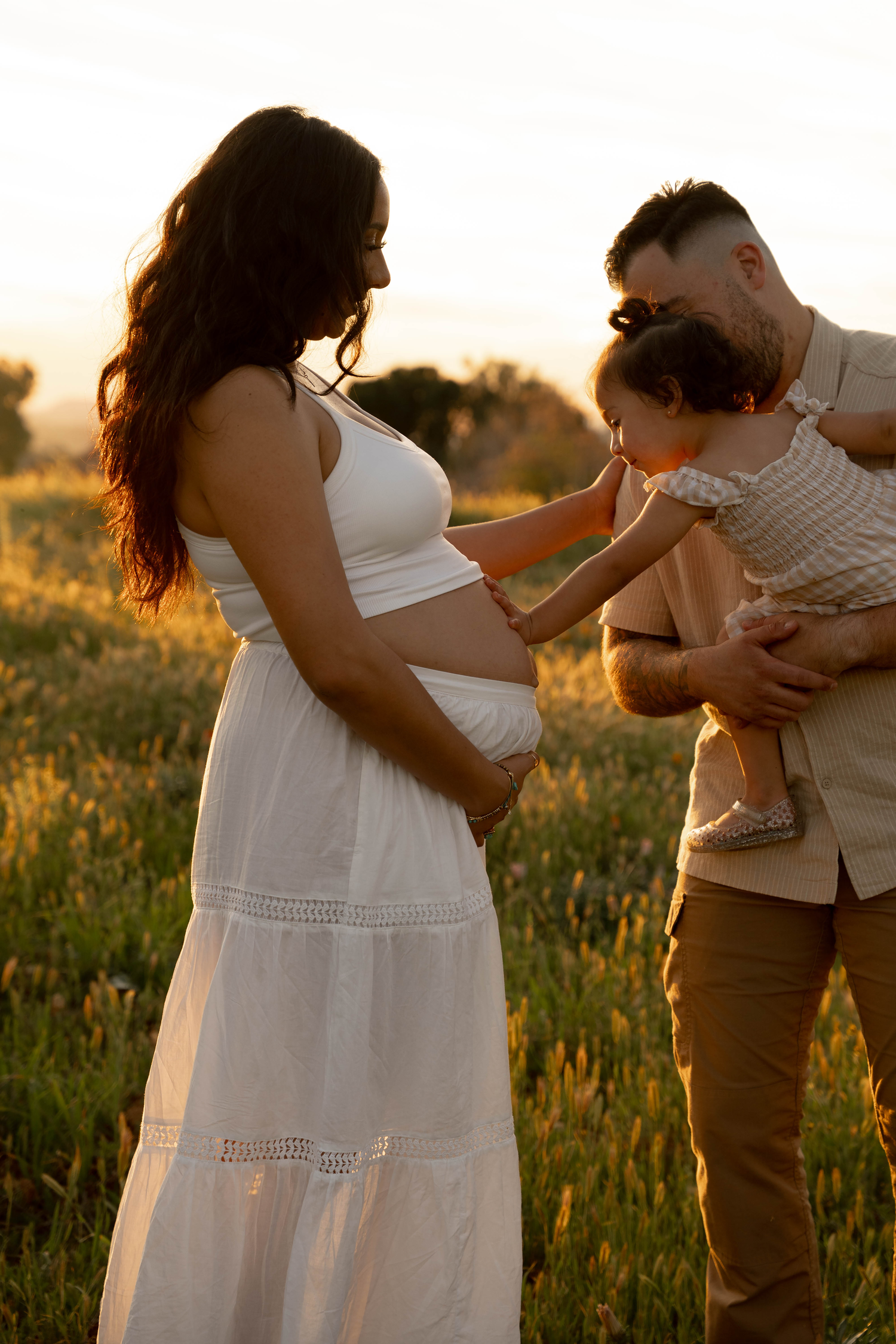 Pregnant woman in white dress with husband holding toddler touching baby bump at golden hour in field.