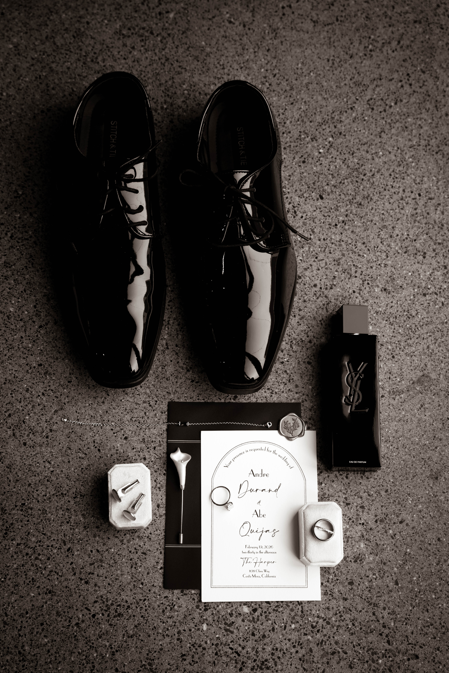 Black and white flat lay of groom accessories including patent leather shoes, cufflinks, cologne, rings, and wedding invitation.