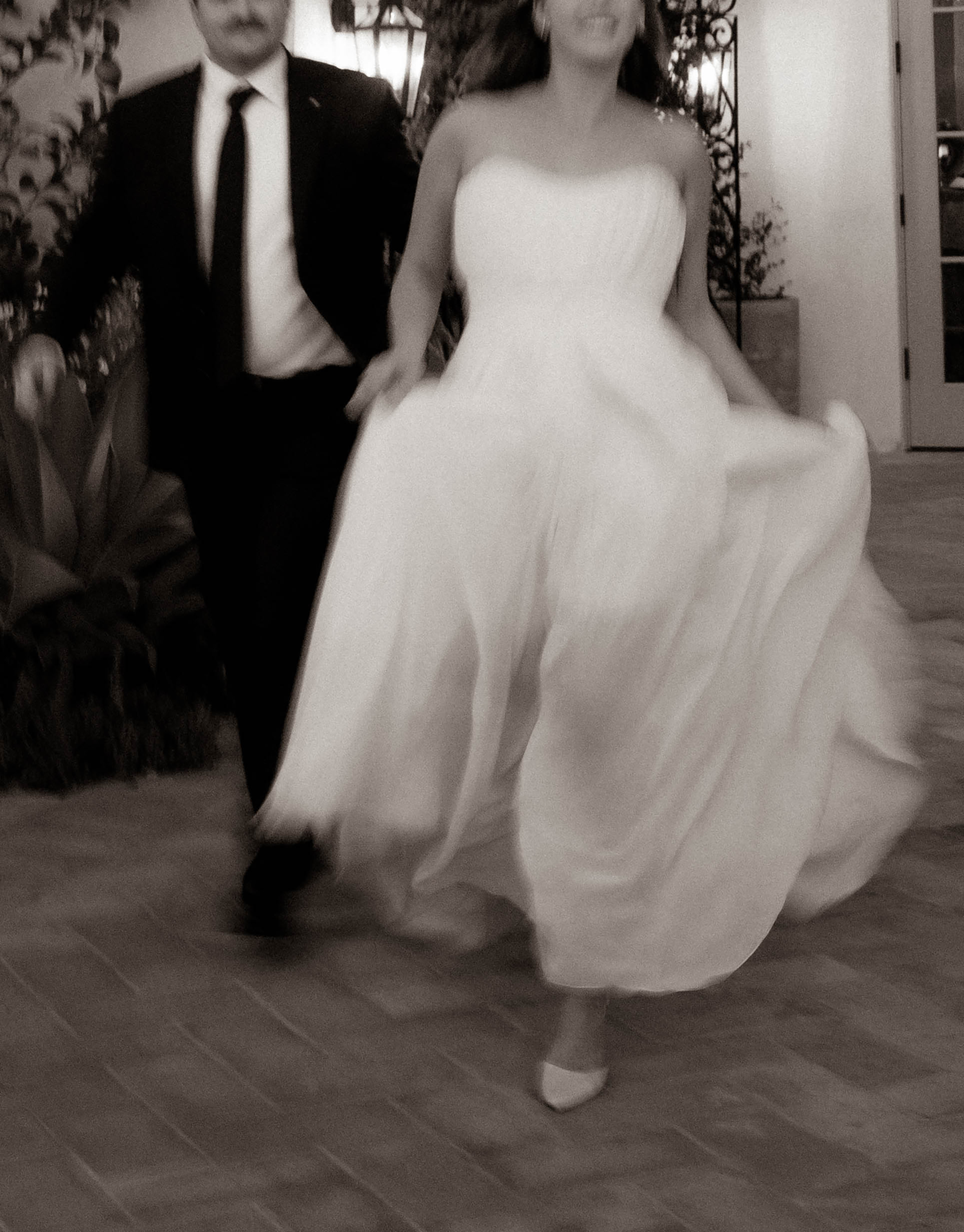 Bride in flowing white gown and groom in black suit walking together at wedding reception in black and white photo.