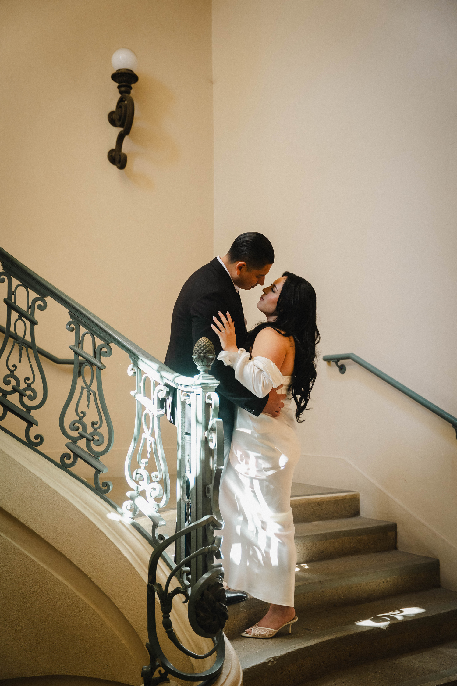 A couple shares a romantic kiss on an elegant staircase, bride in white gown and groom in black suit.