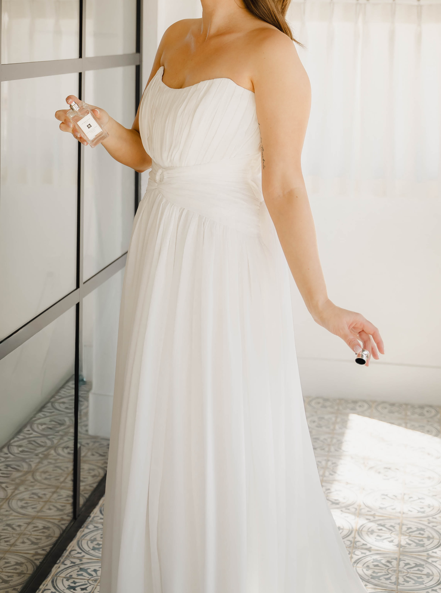 Bride in strapless white chiffon wedding gown holding perfume bottle, standing near mirror on patterned tile floor.