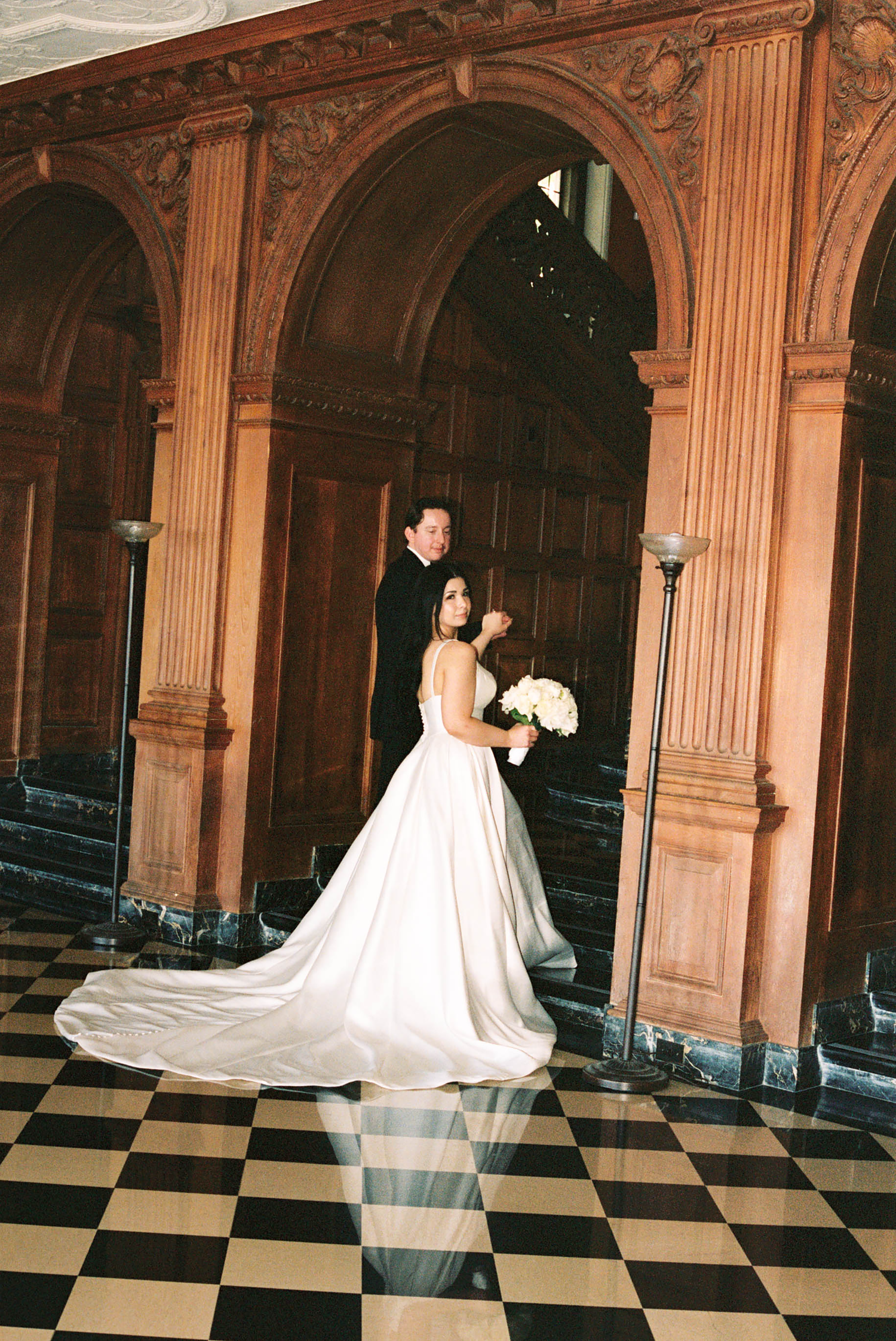 Bride in white ball gown with long train and groom in black tuxedo pose on checkered floor by ornate wooden arches.