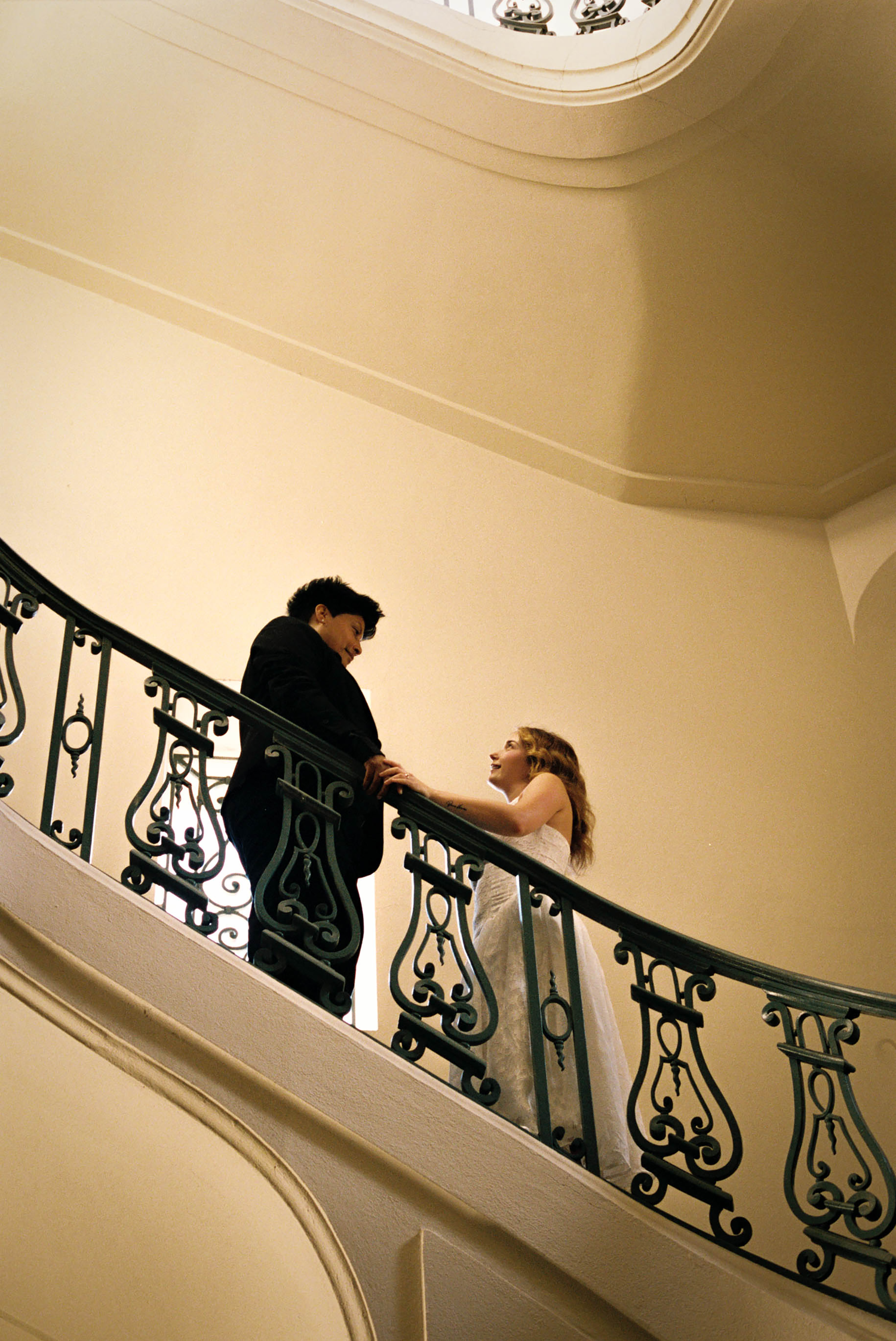 A couple in wedding attire stands on an elegant curved staircase with ornate iron railings in a grand venue.