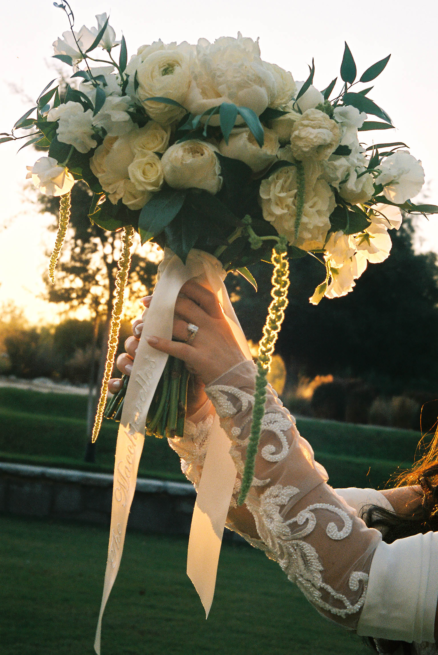 Bride holding white rose wedding bouquet with gold chain accents and ribbon wrap at sunset outdoor ceremony.
