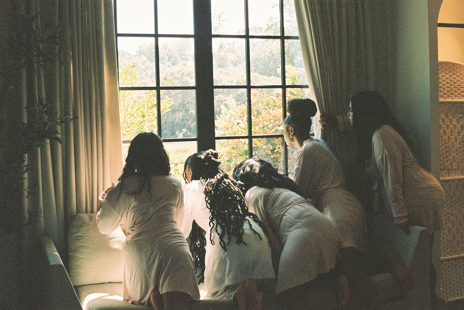 Group of women in white robes gathered together looking out a large window with lush green trees outside.