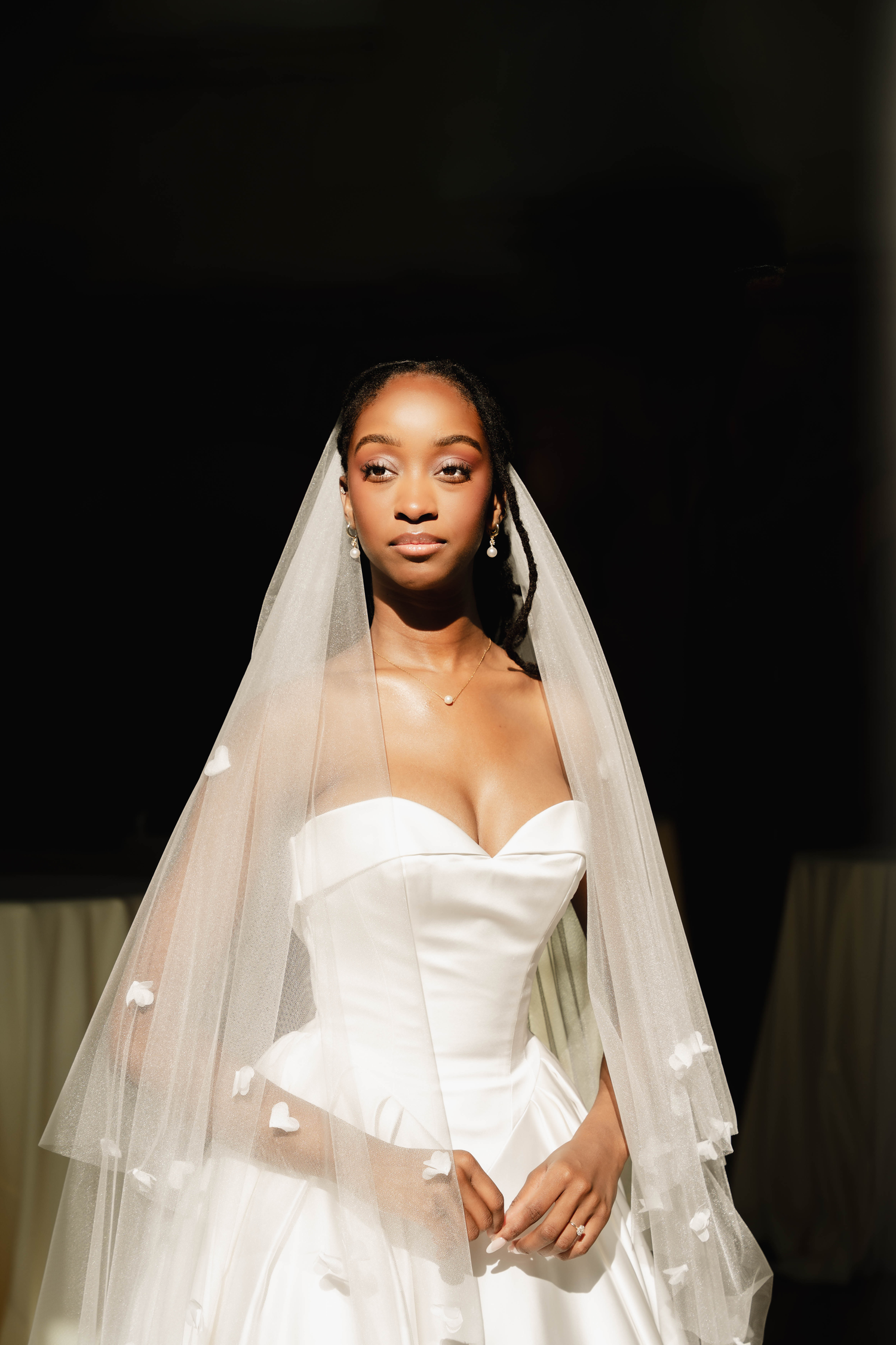 A Black bride in a white strapless ball gown and tiered veil with floral trim poses elegantly against a dark background.