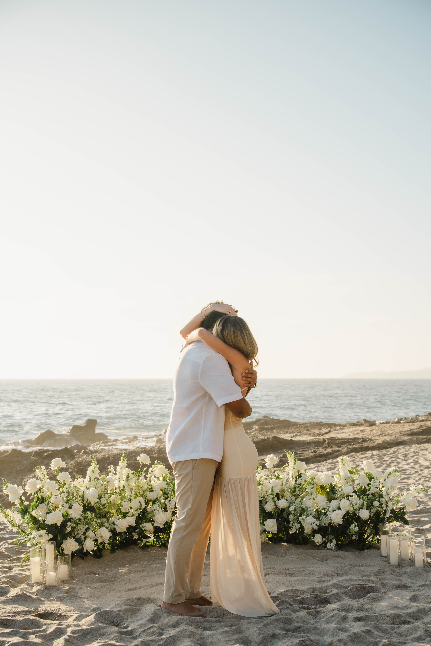 Couple sharing tender beach embrace surrounded by white floral arrangement and candles at sunset.