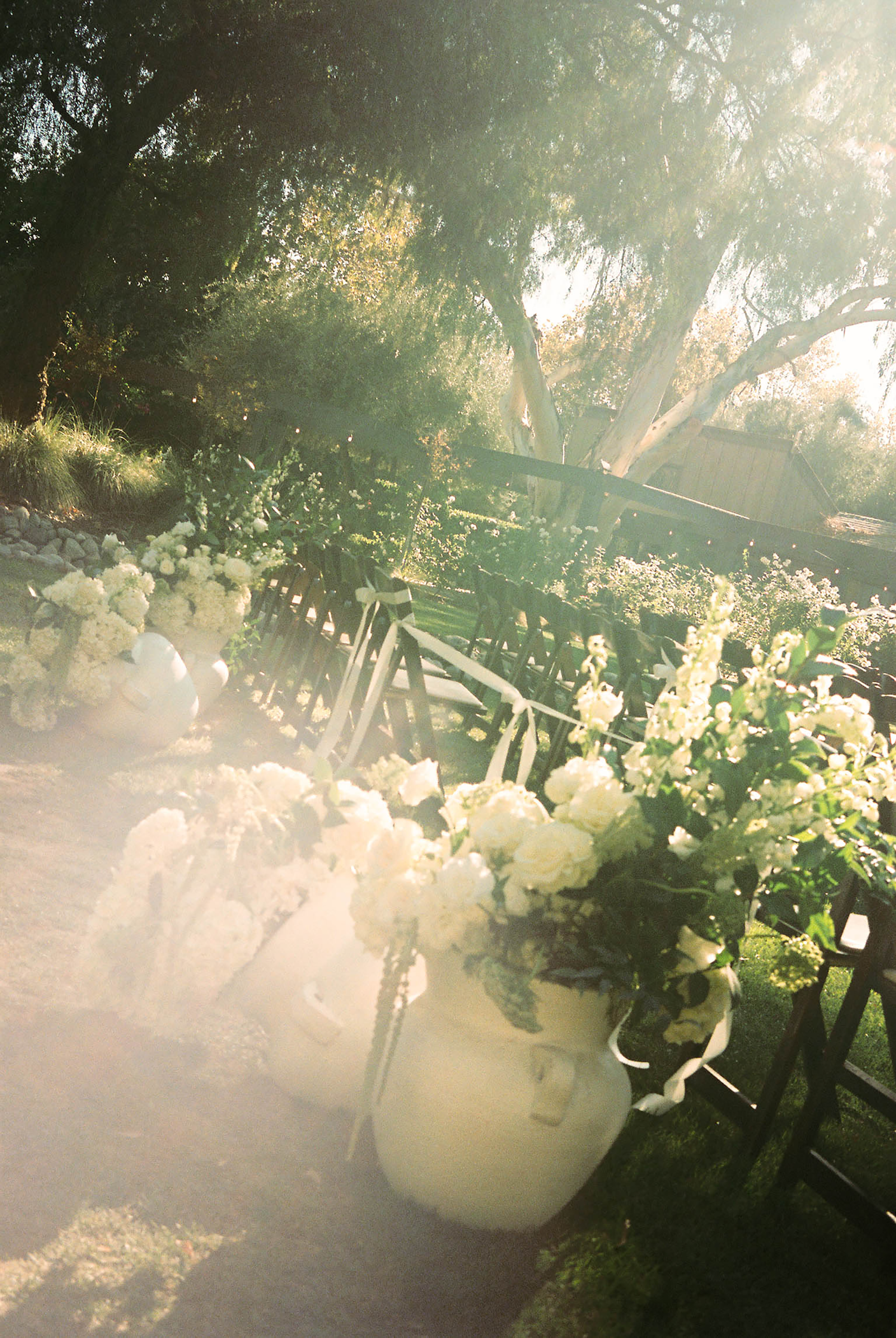 Outdoor wedding ceremony aisle with white floral arrangements in buckets, green chairs, and sunlit trees in the background.