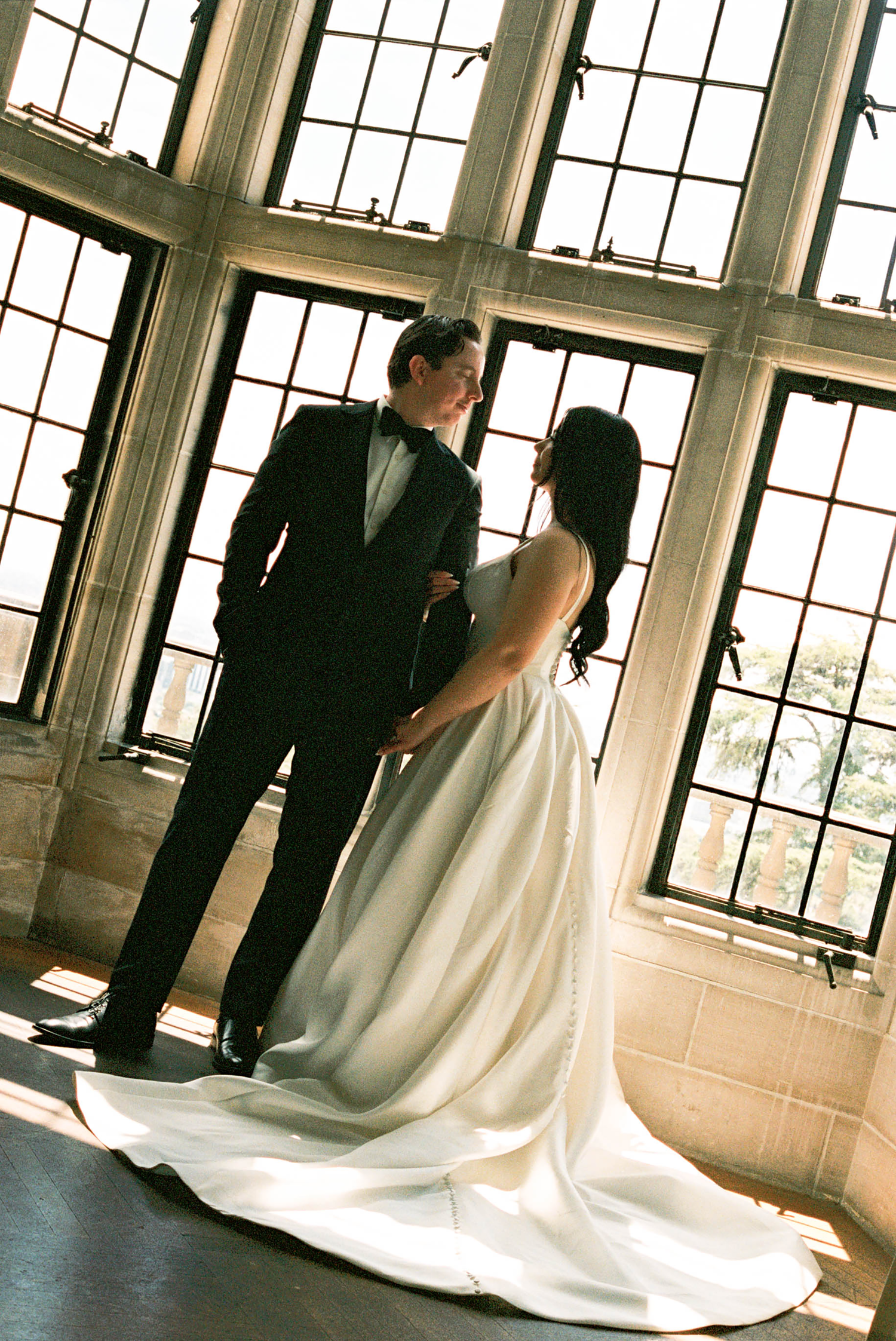 A bride and groom share an intimate moment in front of large windows, bride in a flowing white gown, groom in a black tuxedo.