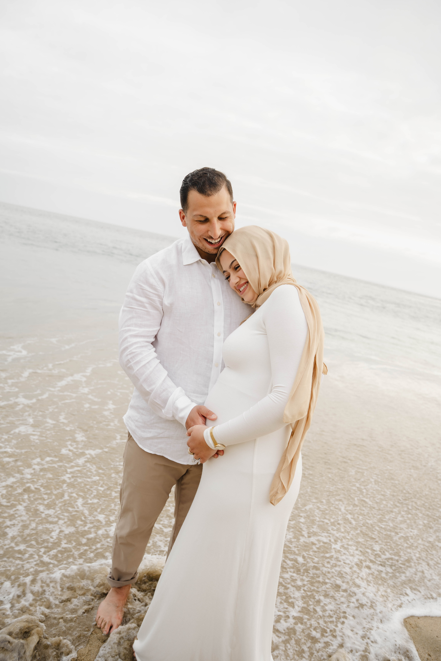 A couple embraces on a beach, the woman in a white dress and hijab leaning on a smiling man in a white shirt.