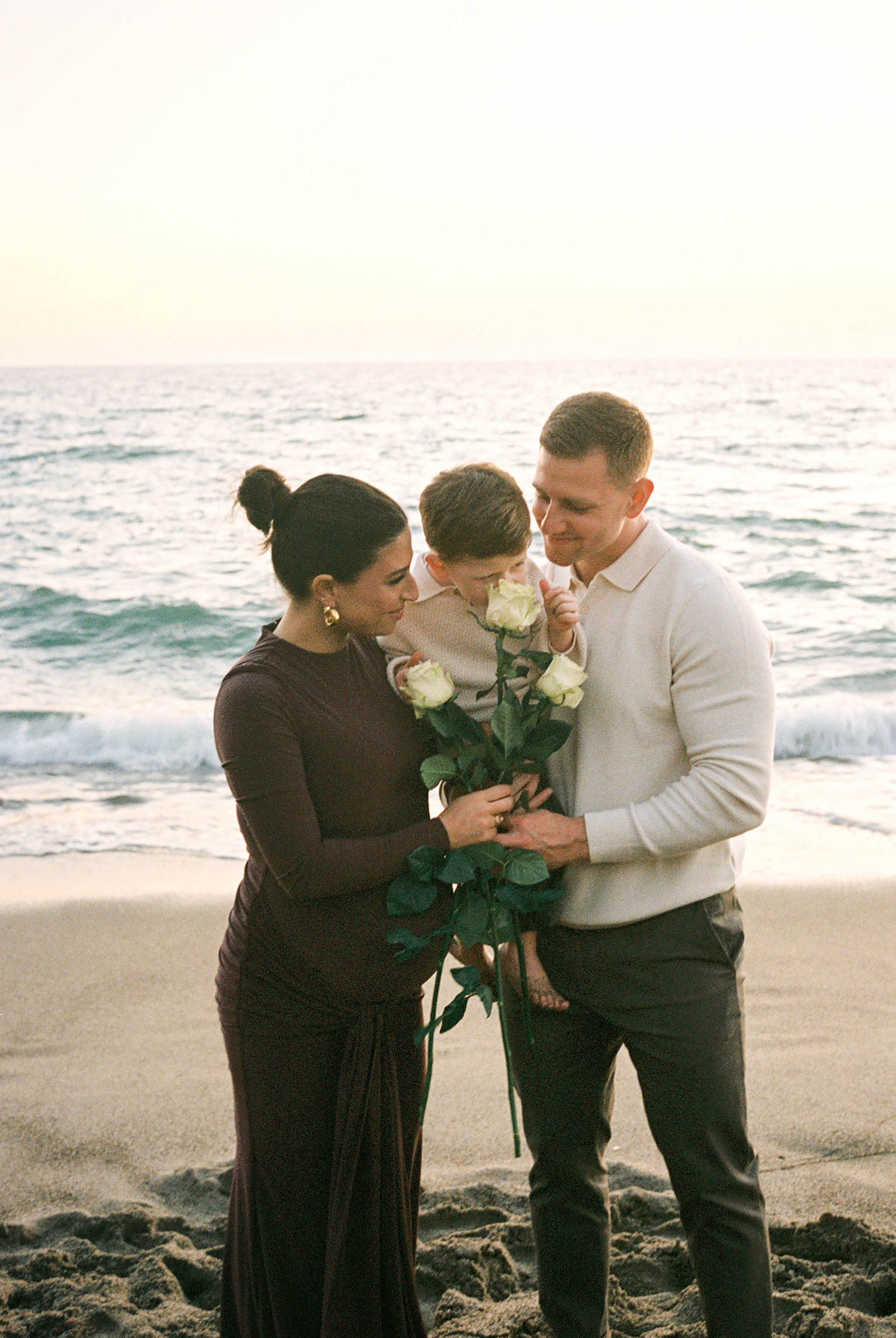 Family of three sharing white roses on a sandy beach at sunset, ocean waves in the background.
