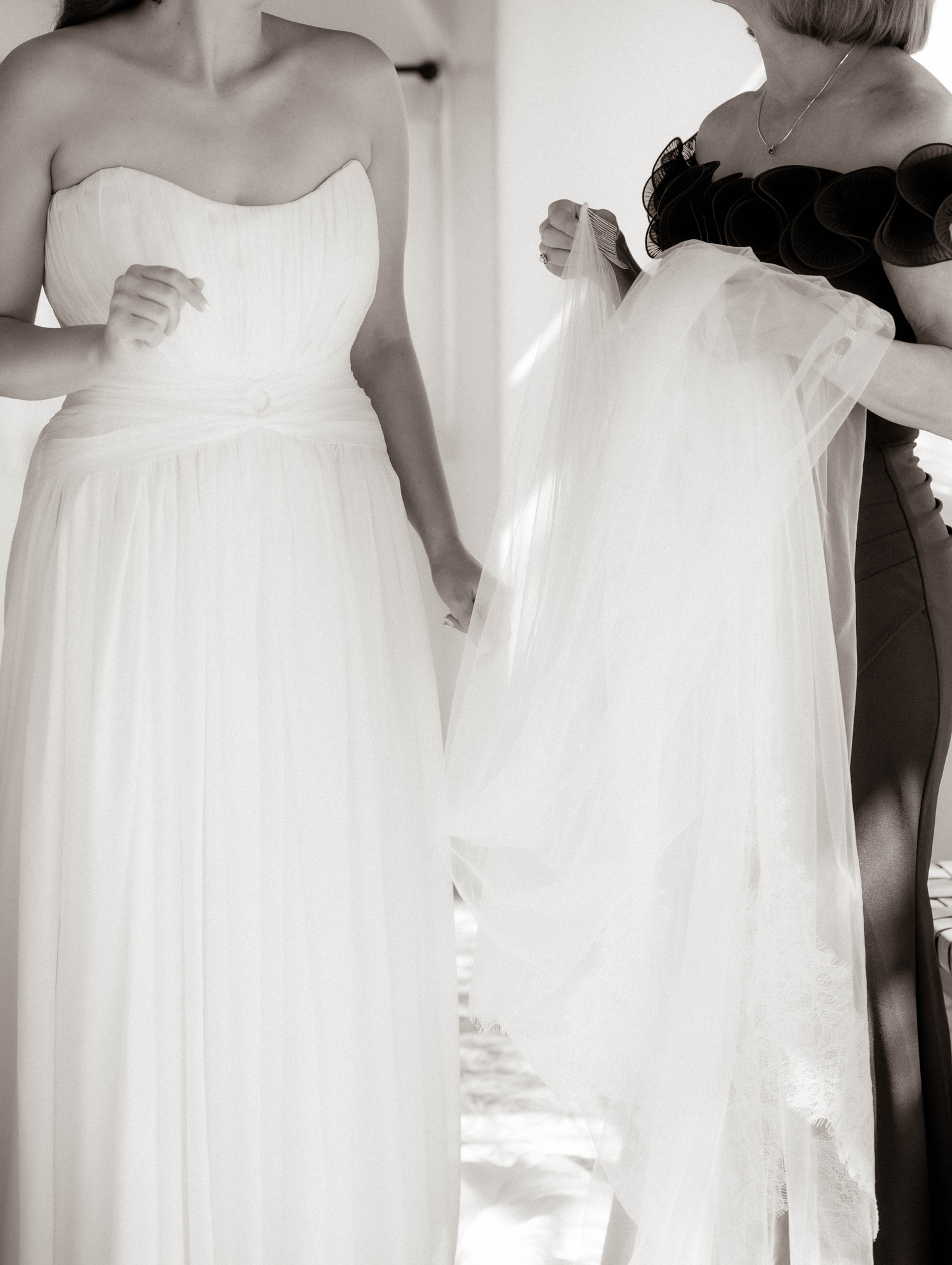Black and white photo of bride in strapless chiffon gown being helped with veil by bridesmaid.